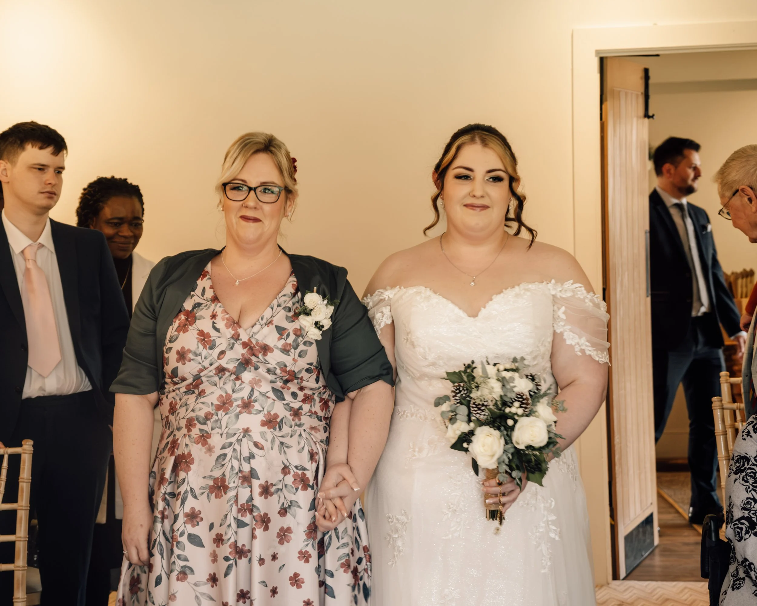 A bride holding a bouquet standing next to a woman at a wedding ceremony, with other guests in the background.