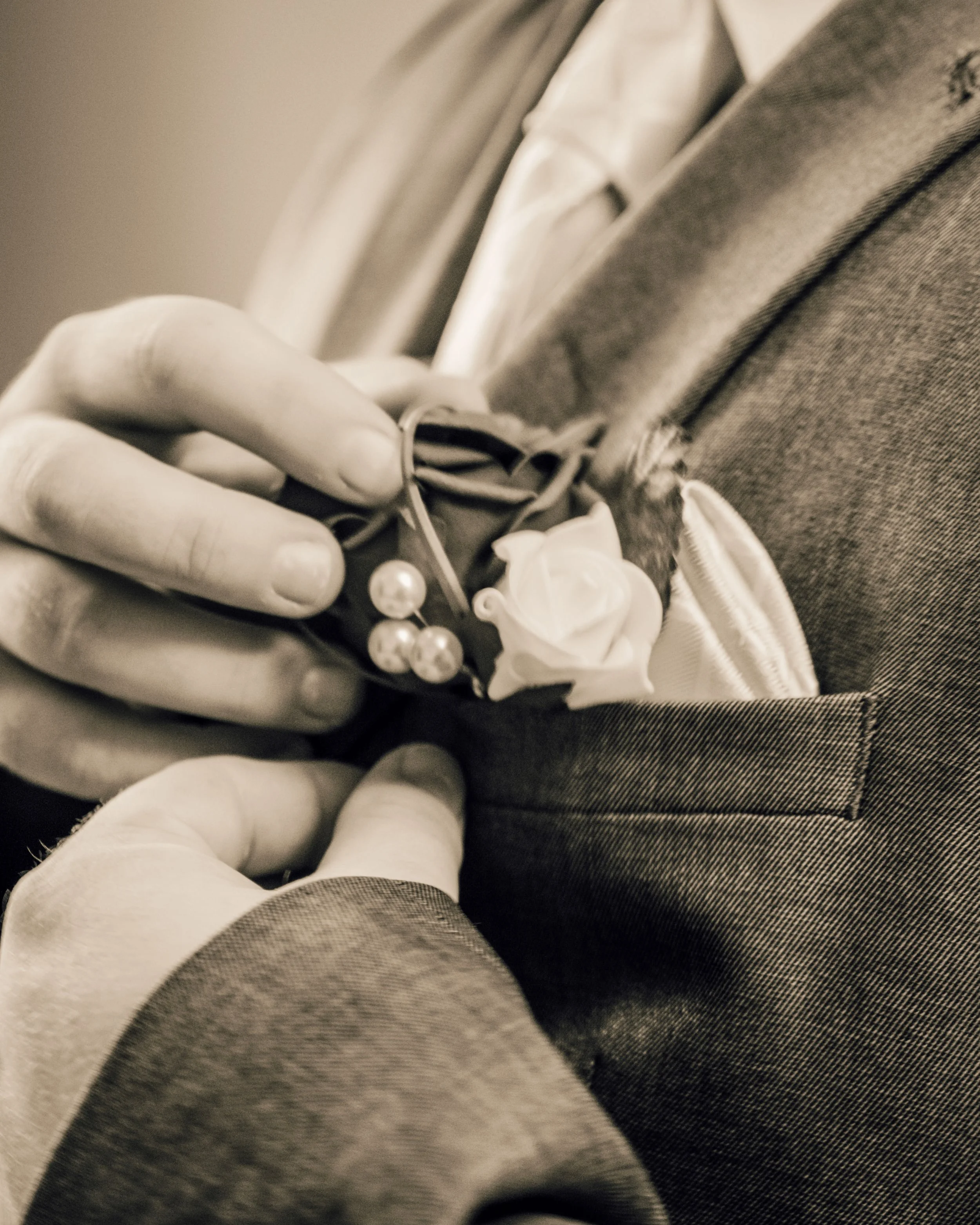 Close-up of a hand adjusting a boutonniere on a lapel, with a focus on the flower and ribbon, blurred background.