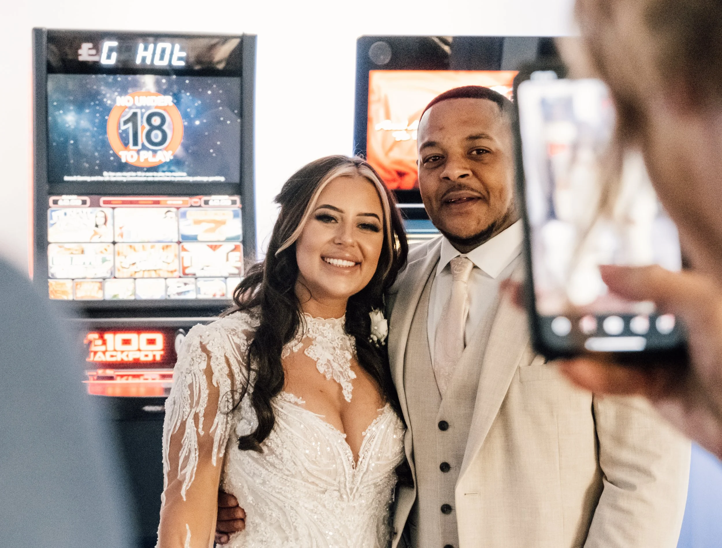 A newlywed couple taking a selfie in front of slot machines at a casino. The bride is smiling, wearing a white lace wedding dress, and the groom is dressed in a light-colored suit with a white shirt and tie. The background features electronic gaming 