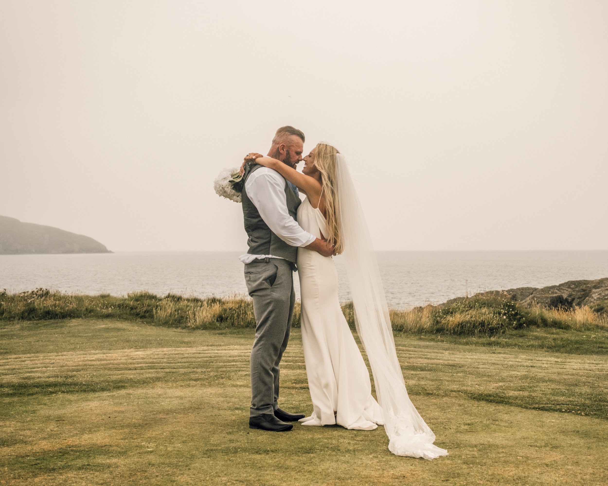 A groom and bride embrace each other lovingly on a grassy field, with water and hills in the background, during their wedding celebration.