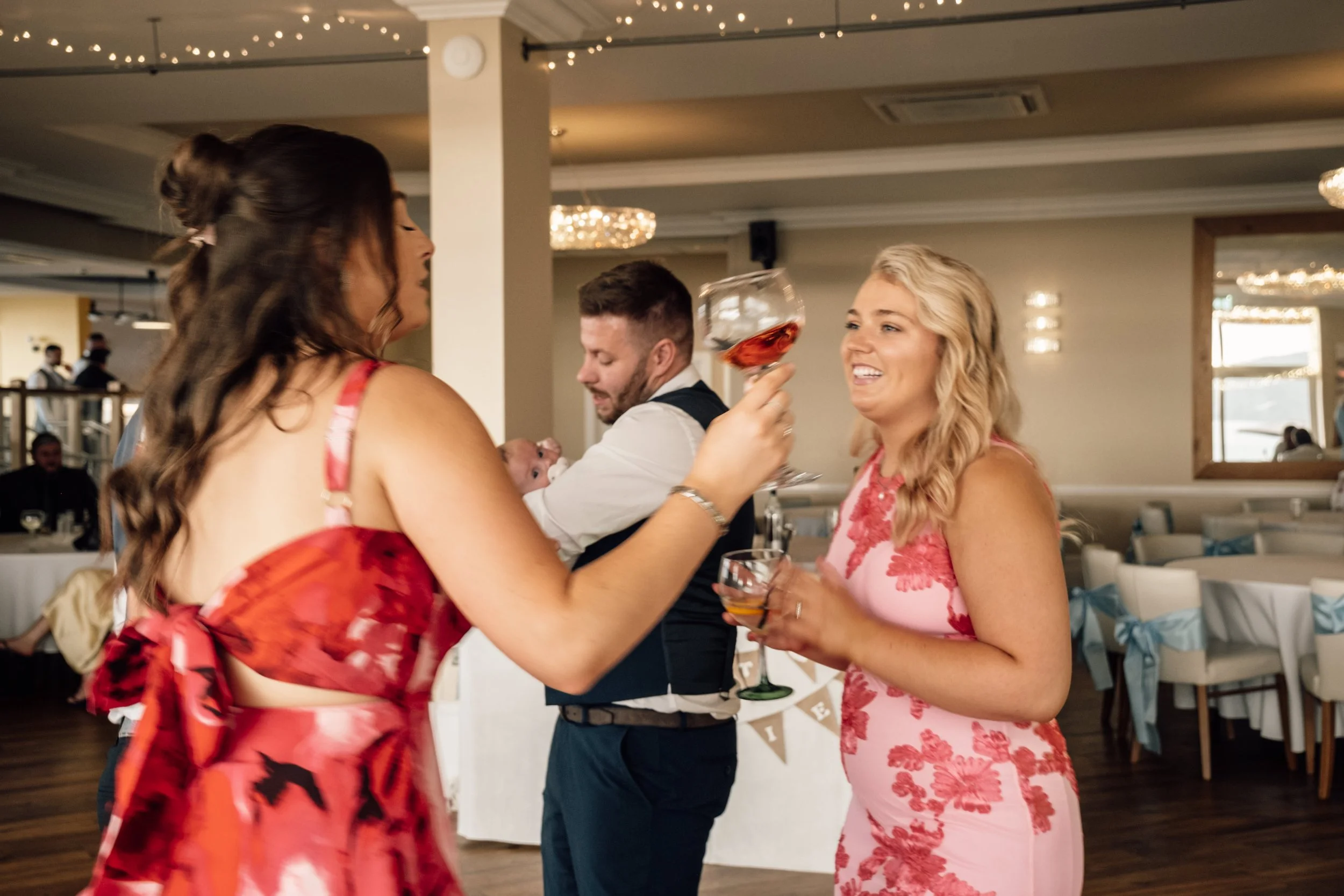 Three women at a celebration or party, with one holding a large glass of rosé wine, smiling and engaging with each other, while a man in the background holds a baby, smiling, in a decorated indoor venue.
