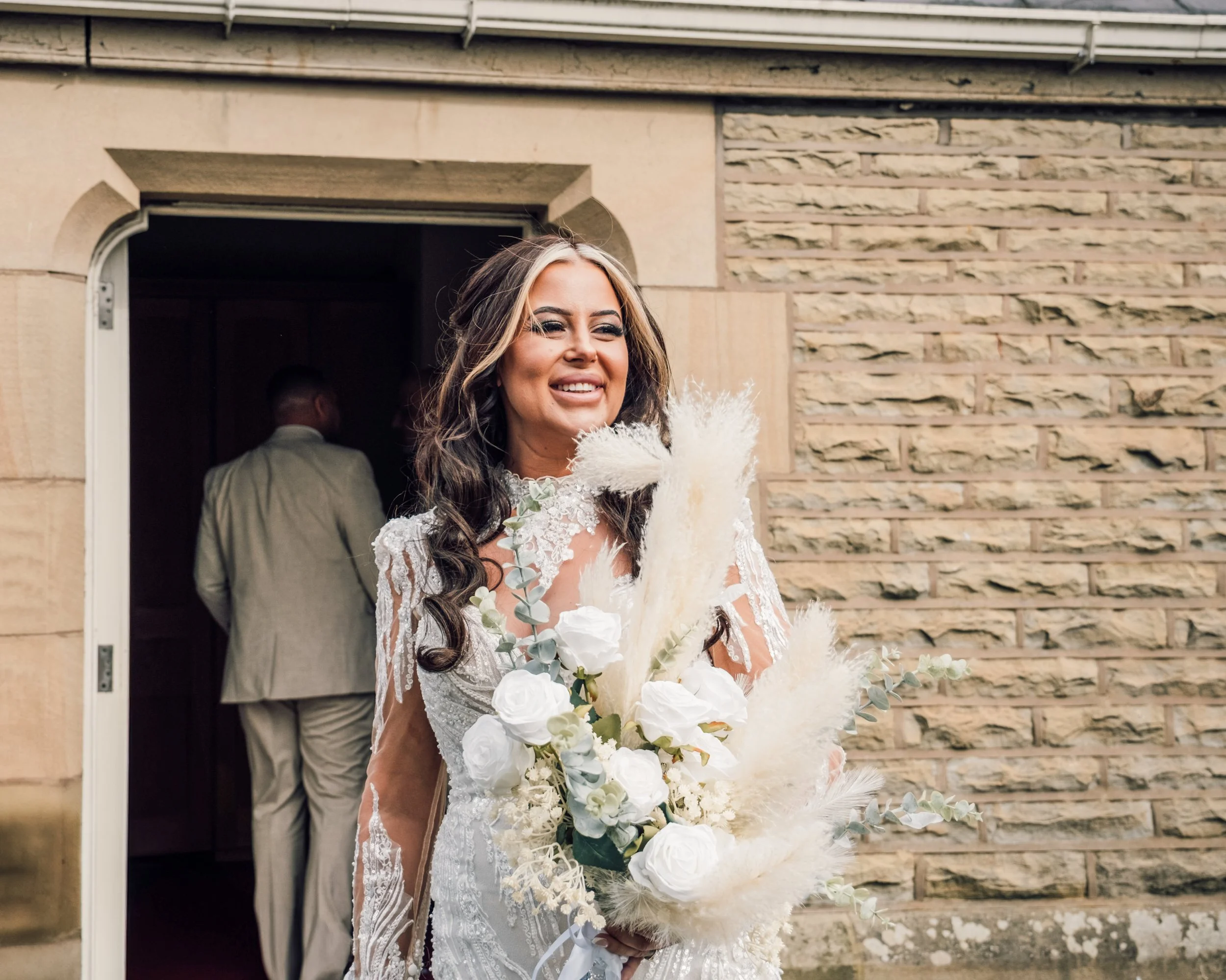 A smiling bride with long dark hair, wearing a lace wedding gown, holding a bouquet of white roses and eucalyptus, standing in front of a brick wall and a doorway.