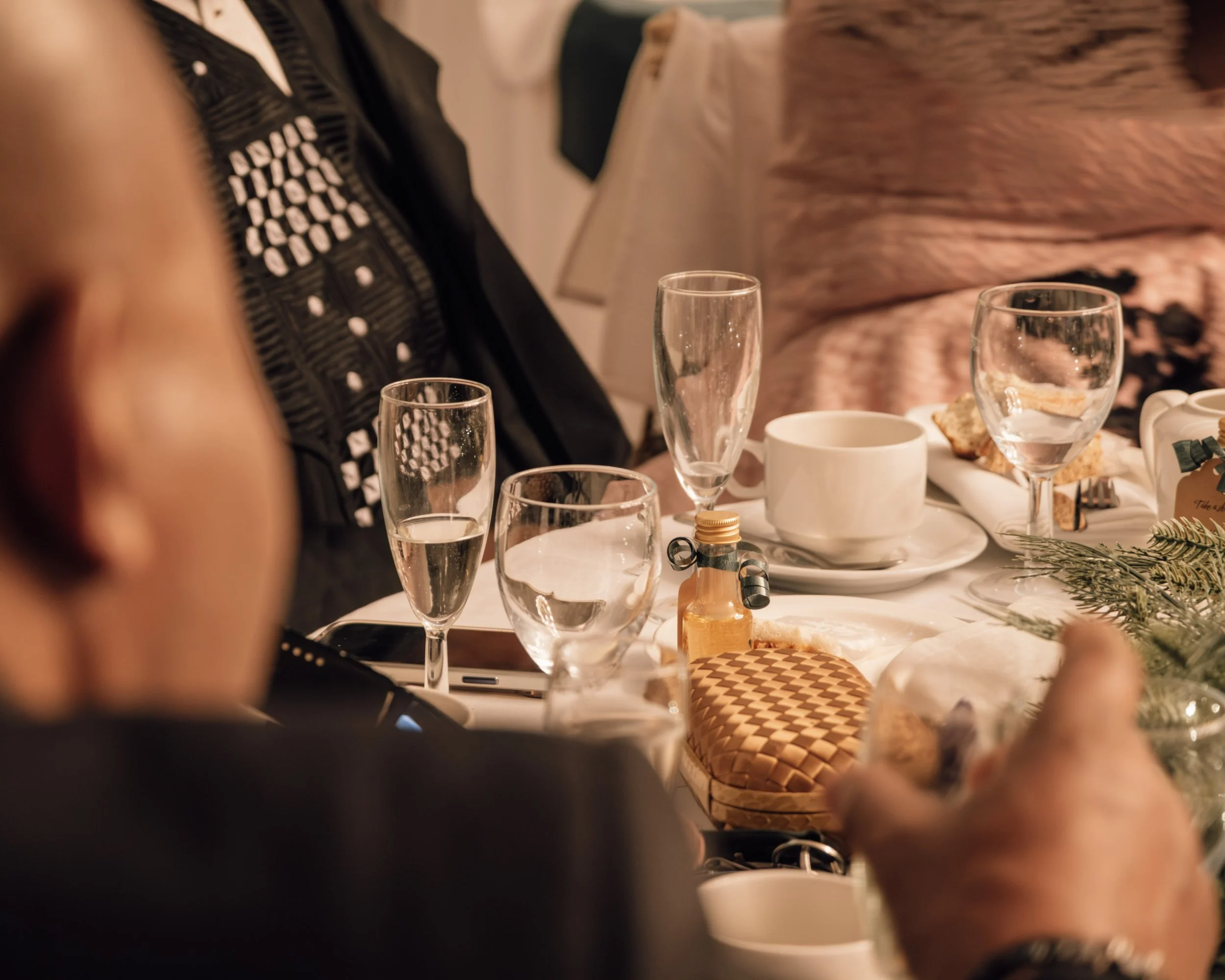 Part of a dining table setup with empty wine glasses, cups, small plates, and assorted food items, with blurred human figures in the background.