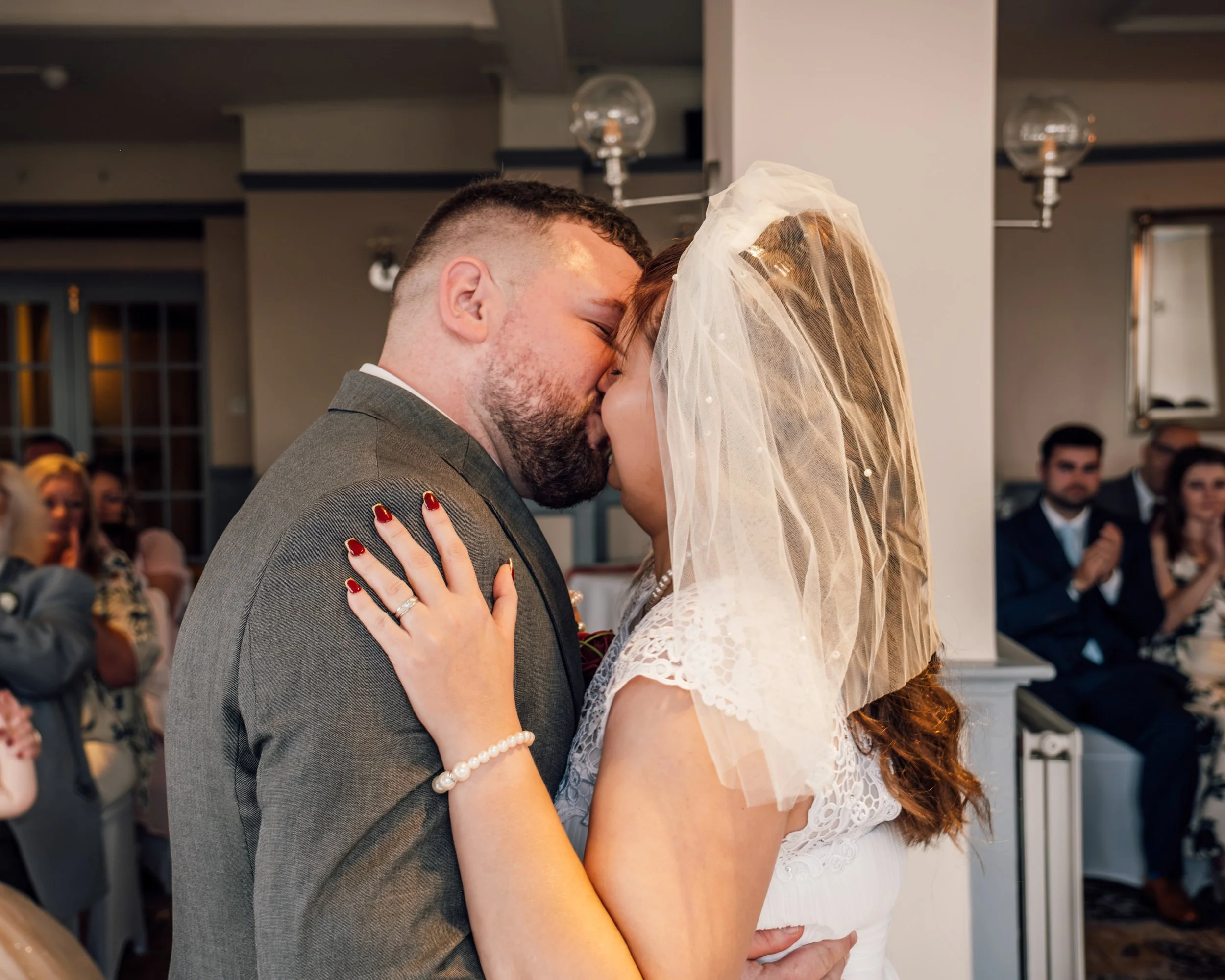 A couple kissing during a wedding ceremony, with the groom wearing a gray suit and the bride in a white lace dress and veil, surrounded by wedding guests.