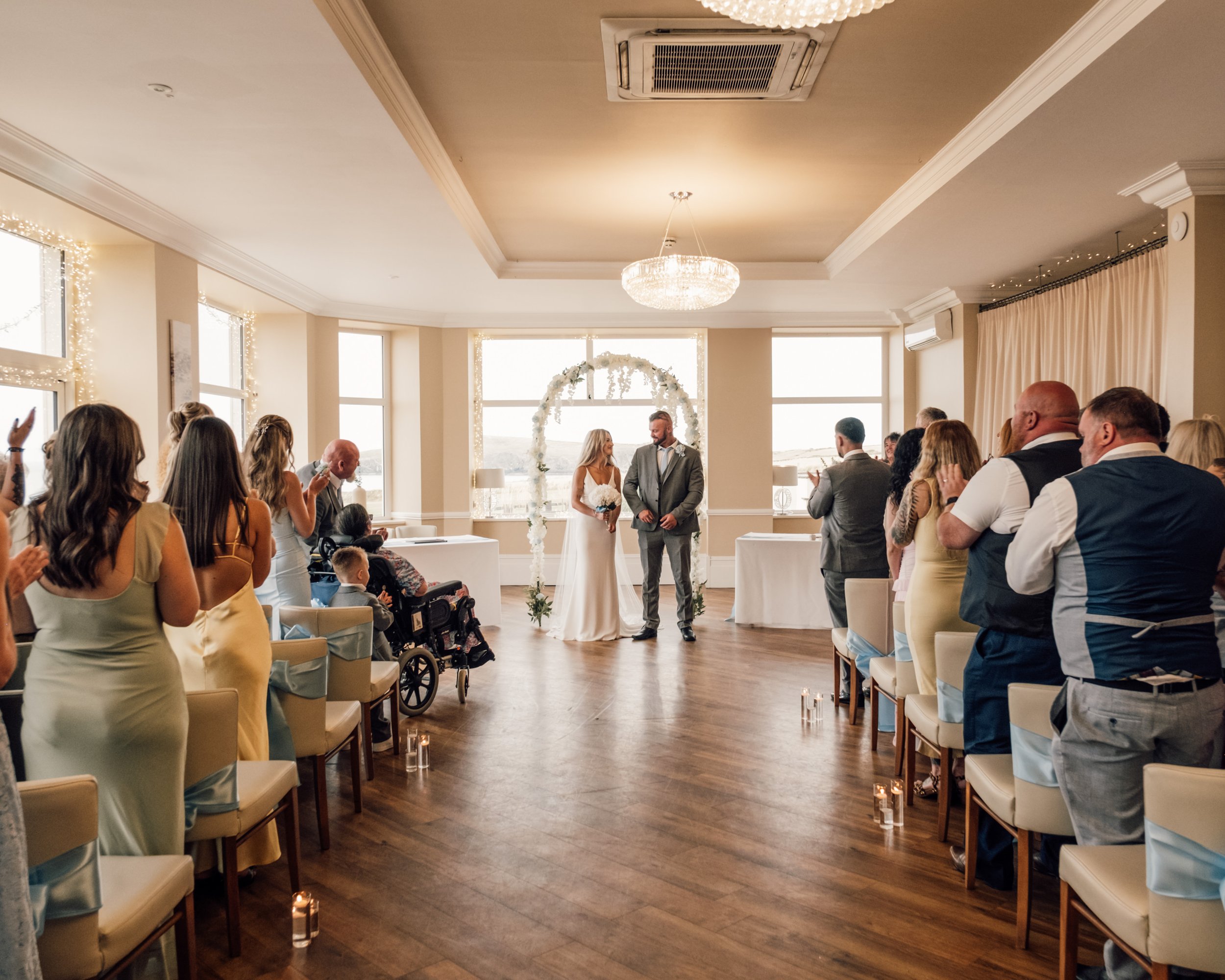 A wedding ceremony taking place indoors, with a bride and groom standing under a floral arch, surrounded by family and friends. The room has large windows, a chandelier hanging from the ceiling, and candlelit decorations along the aisle.