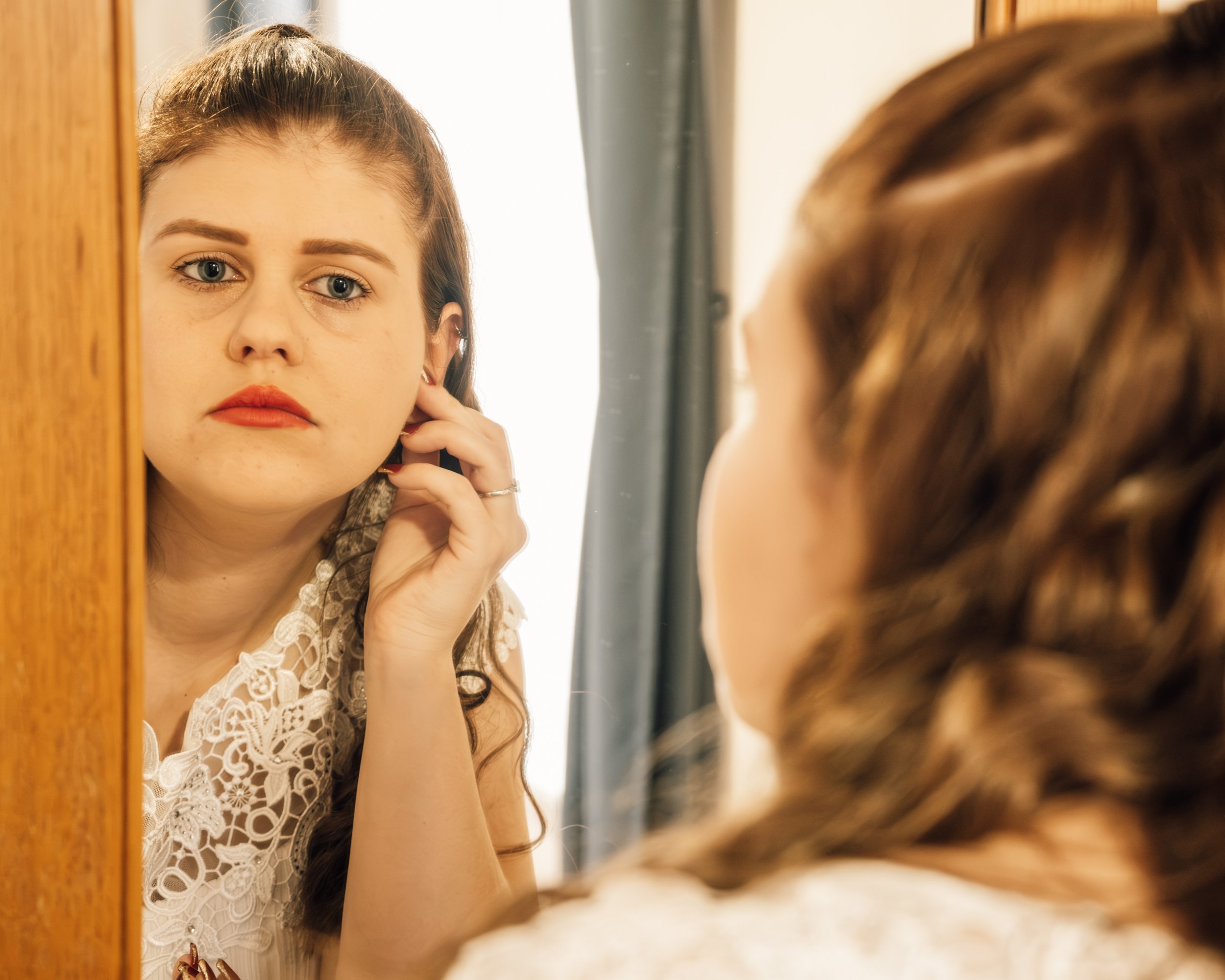 A young woman with blue eyes and light brown curly hair looking at herself in a mirror, touching her ear with one hand, wearing a white lace blouse, standing indoors near a wooden surface and curtain.