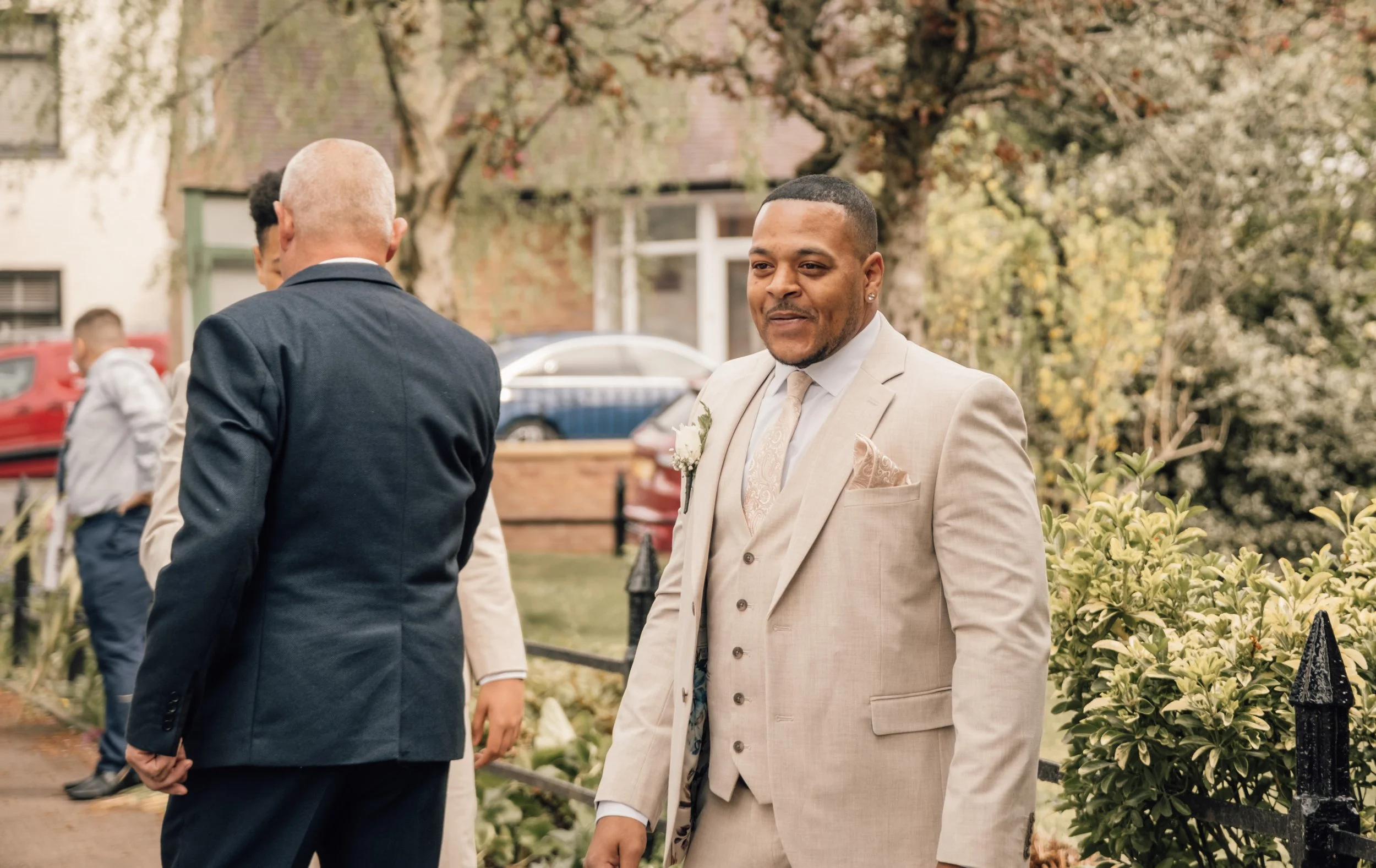 A man in a beige suit with a boutonniere, standing outdoors on a sunny day, smiling and looking at someone off-camera, with other people visible in the background near trees and a brick building.