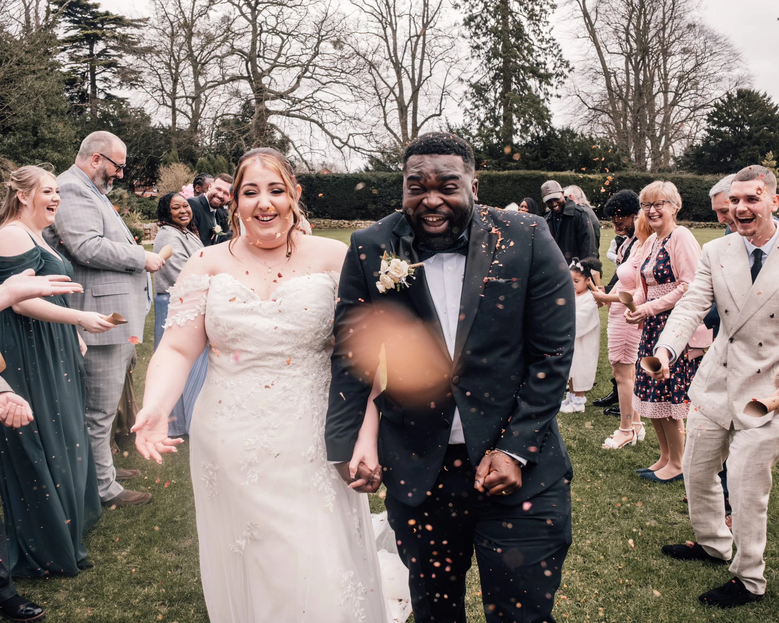 A diverse group of people celebrating a wedding outdoors, with the bride and groom in the center smiling and surrounded by guests throwing confetti.