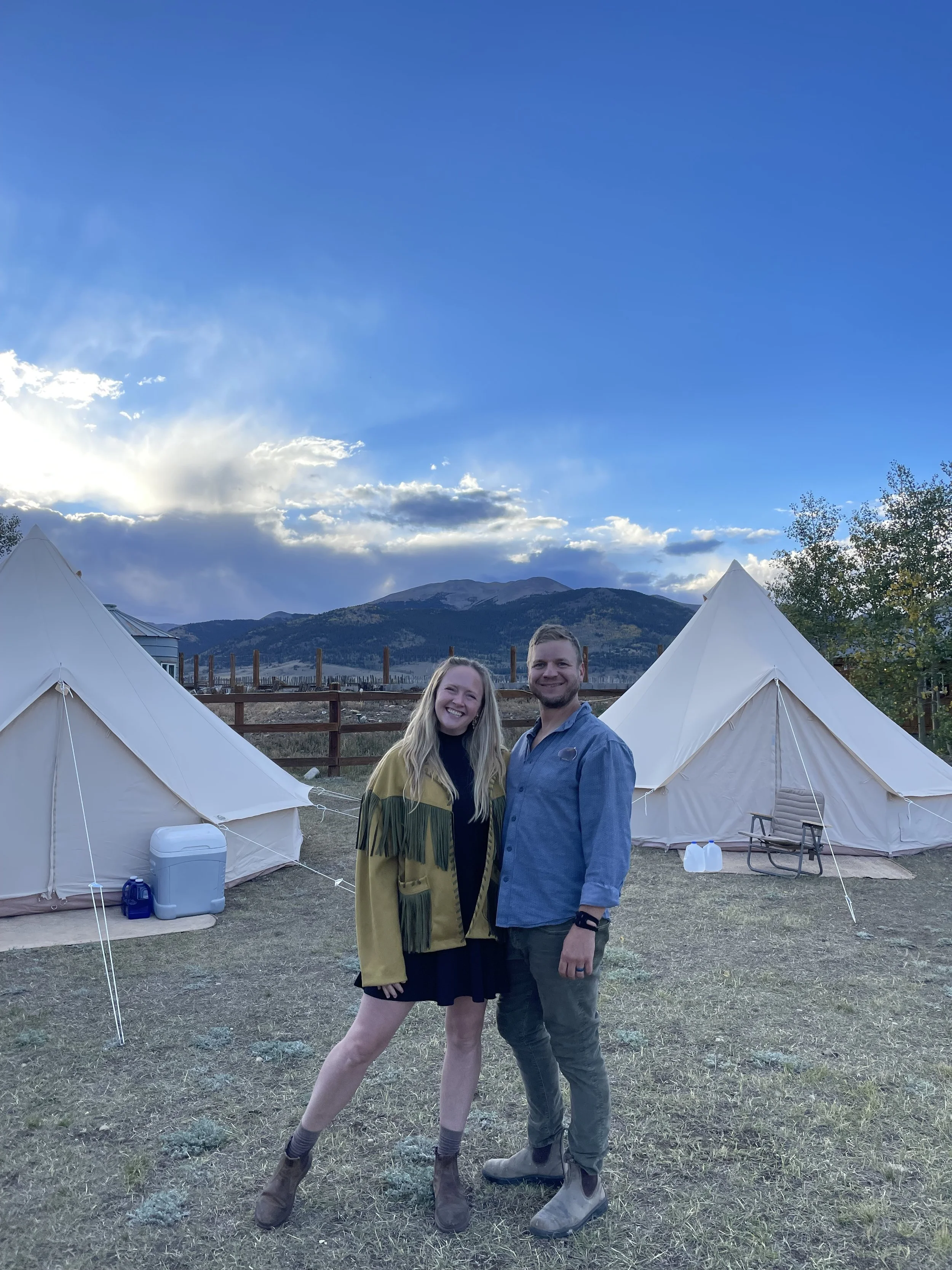 A couple standing outdoors in front of white tents with mountains and partly cloudy skies in the background, smiling and posing for the photo.