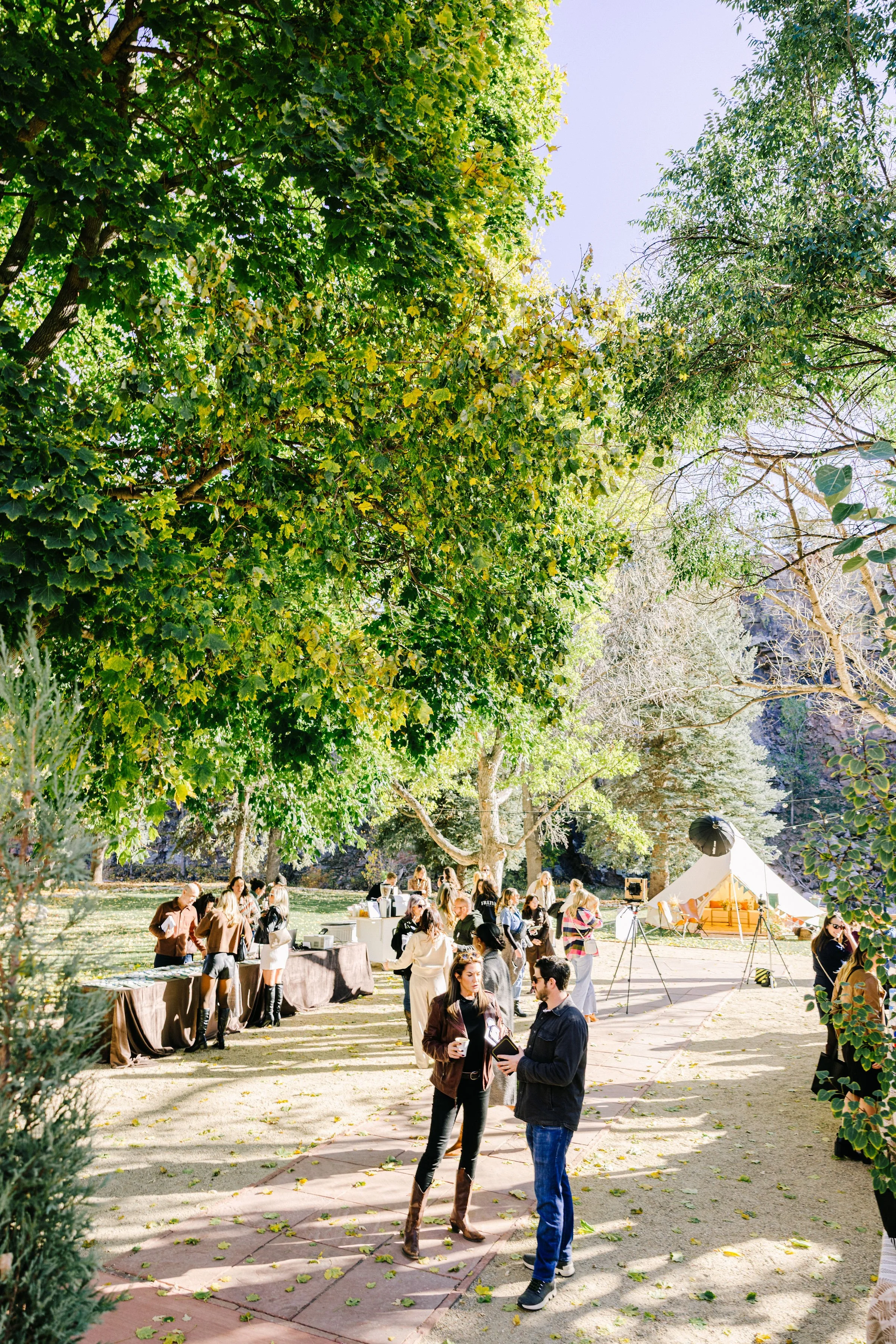 People gather outdoors underneath large green trees on a sunny day, with some standing and chatting and others browsing tables at a market or event. In the background, white tents and equipment set up for a fair or festival.