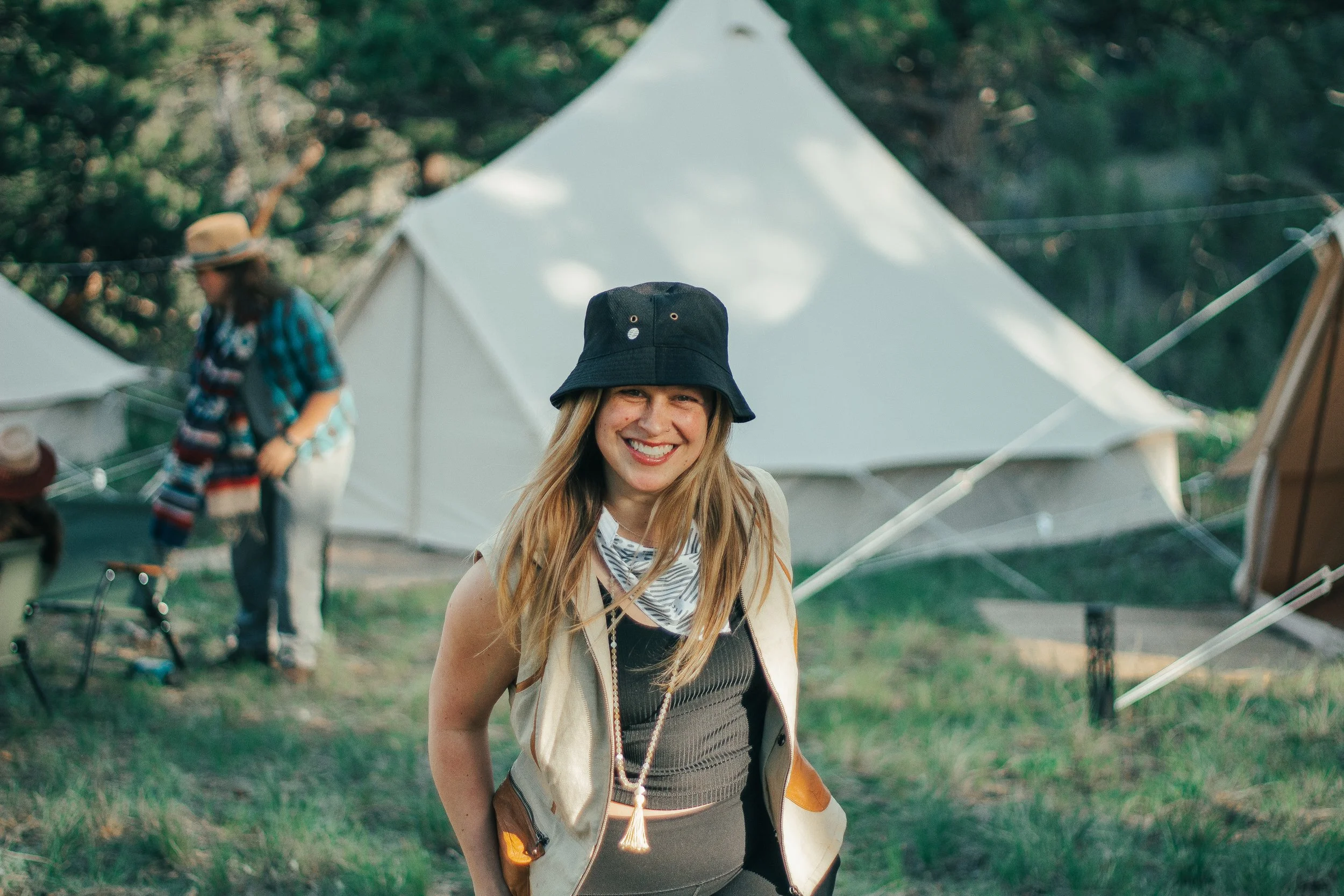 A smiling woman with long blonde hair wearing a black bucket hat, a beige vest, and a black top, standing outdoors near tents at a park or festival.