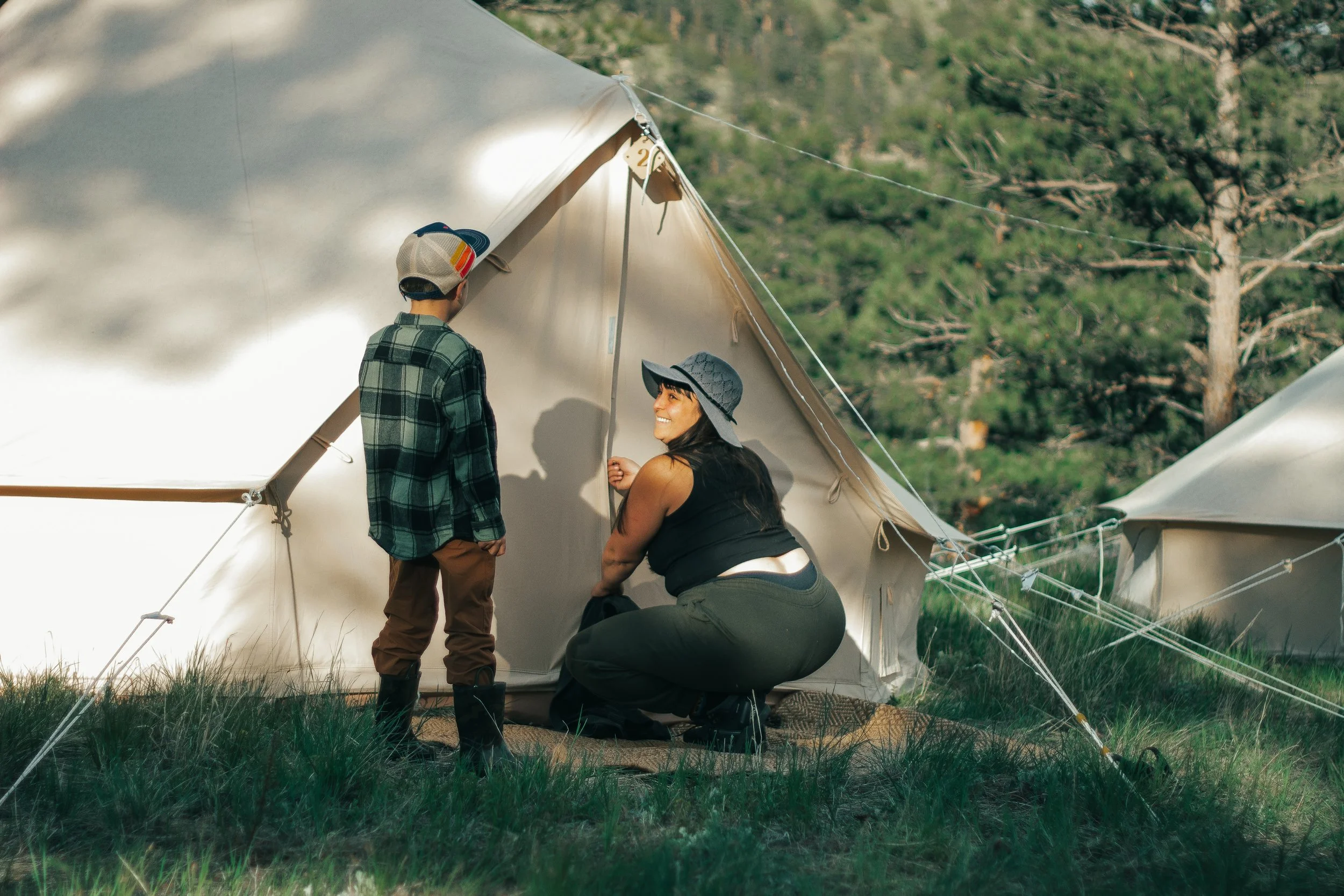 A woman and a young boy inside a beige camping tent outdoors, with a wooded area behind them. The woman is kneeling and smiling, wearing a sun hat, while the boy stands next to her and is facing away, wearing a baseball cap and plaid shirt.