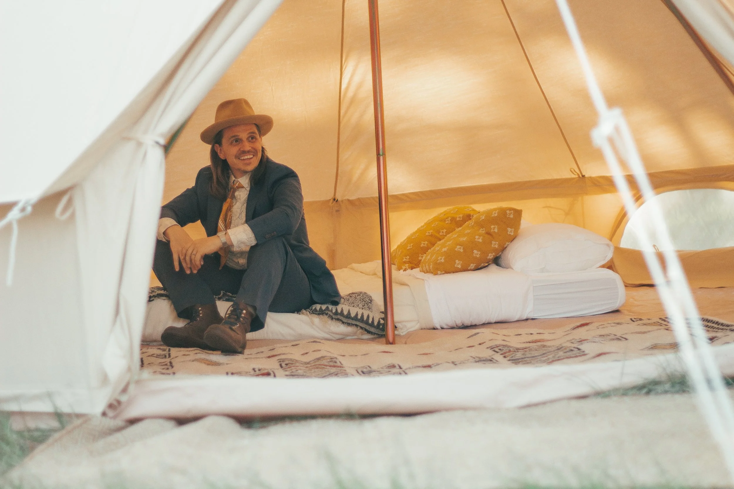 View from inside a canvas tent showing a man in a suit and hat sitting on a bed with yellow pillows, with the tent's structure visible.