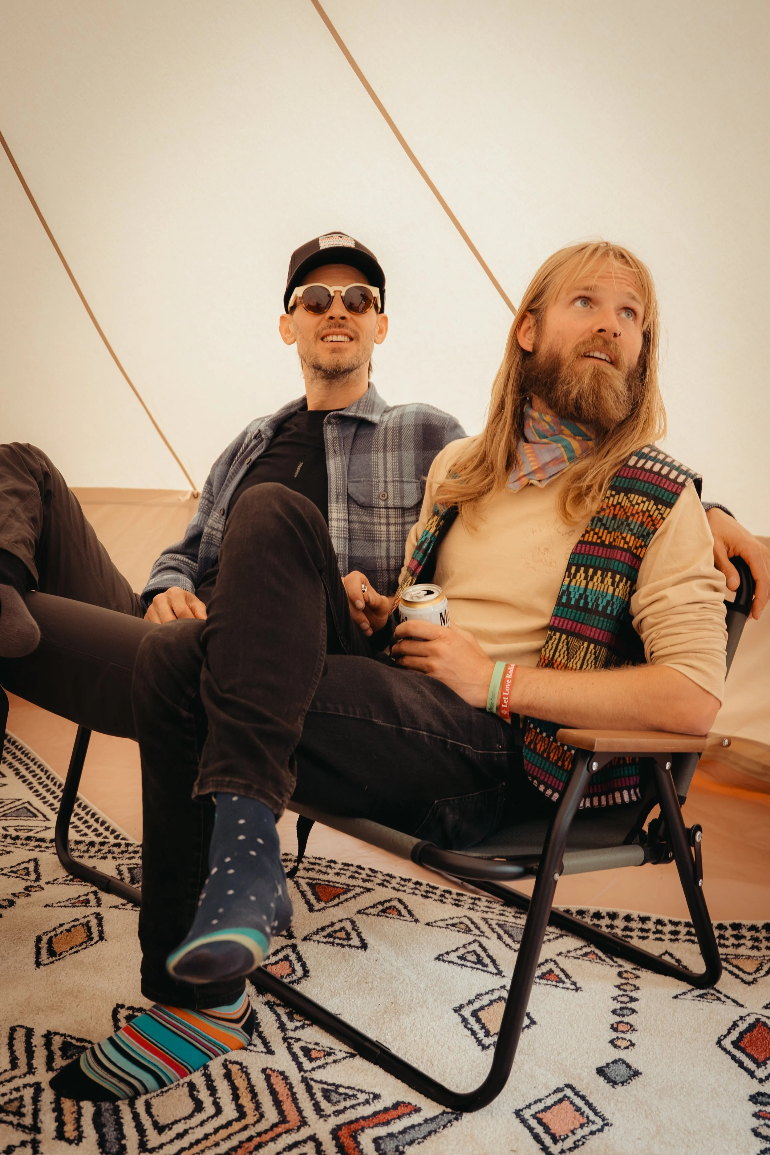 Two men sitting inside a tent, one wearing sunglasses and a hat, the other with long hair and a beard, holding a can of beverage, sitting on a patterned rug.