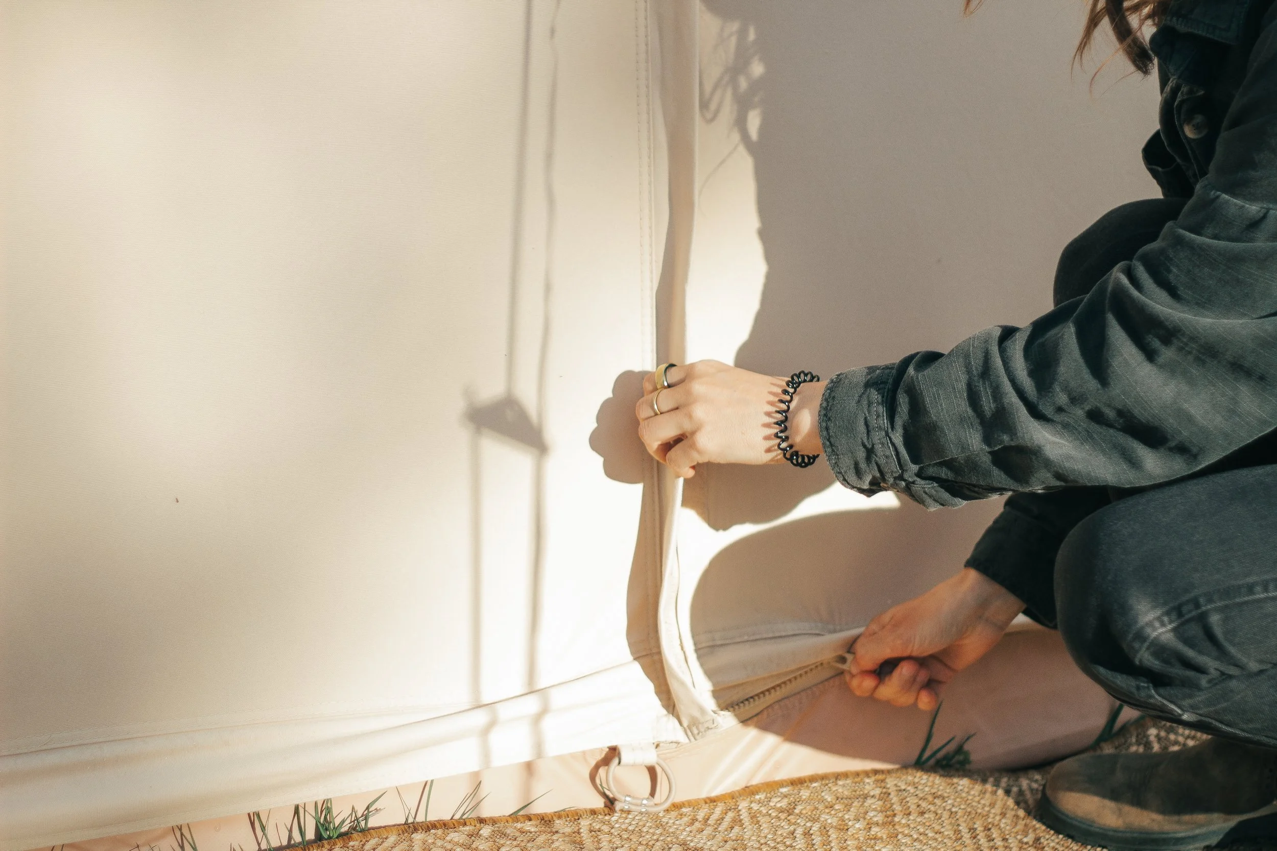 A person wearing a black jacket and multiple rings on their fingers is kneeling down next to a white piece of furniture, possibly a chest or cabinet, with a textured beige and brown rug nearby.