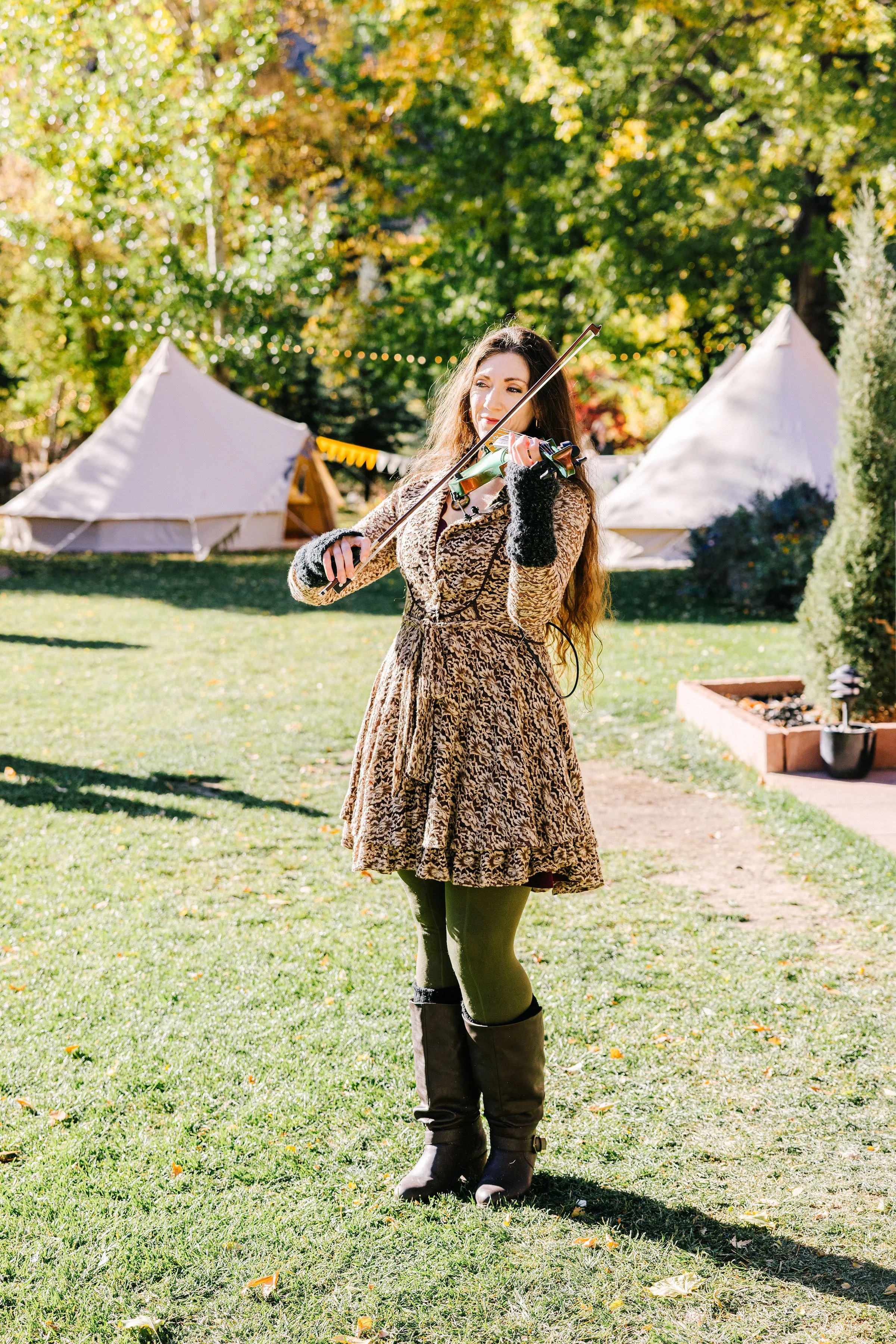 A woman with long hair playing a violin outdoors near white tents on a sunny autumn day.