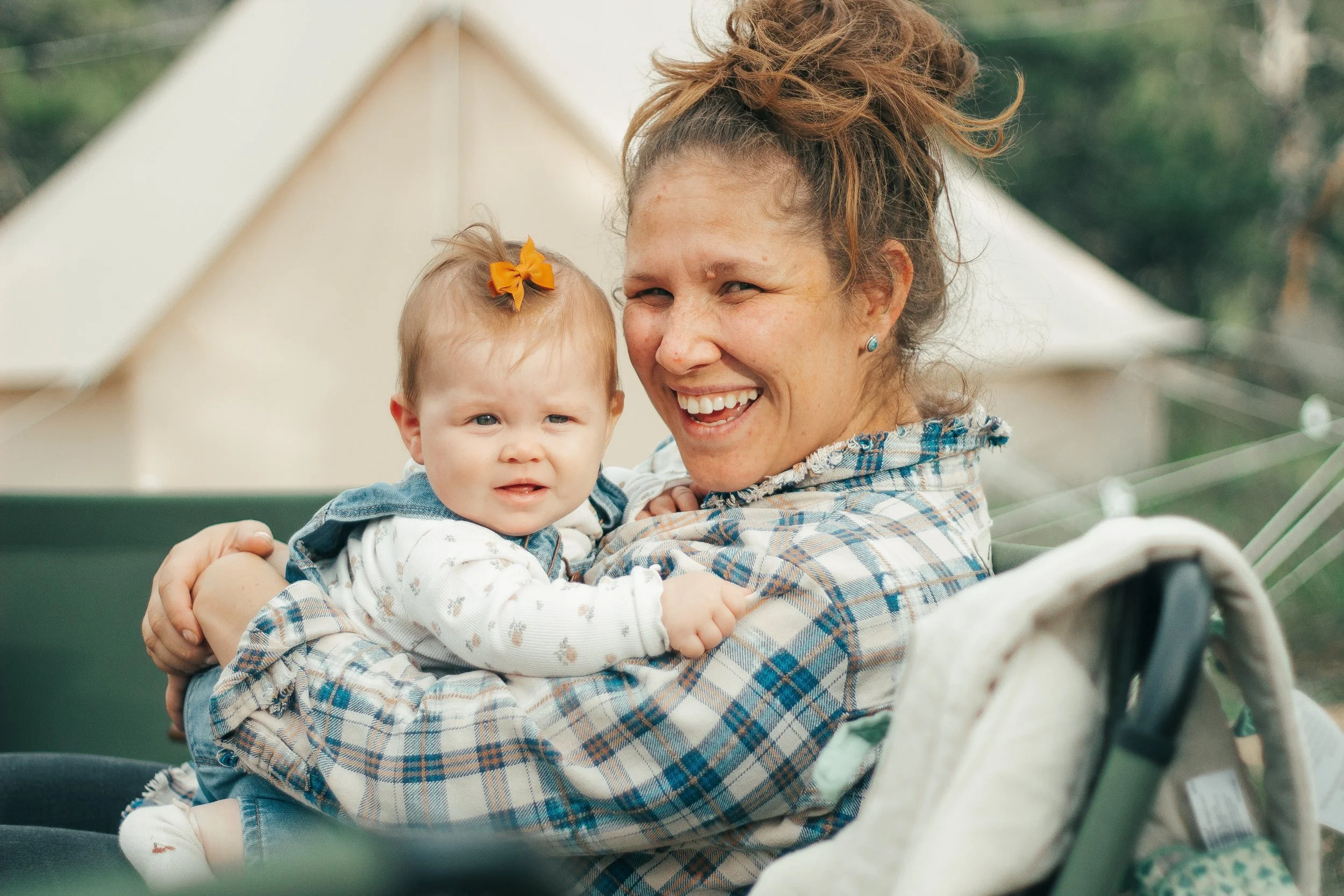 A woman with brown curly hair holding a smiling baby girl with a small orange bow in her hair outdoors near a white tent.