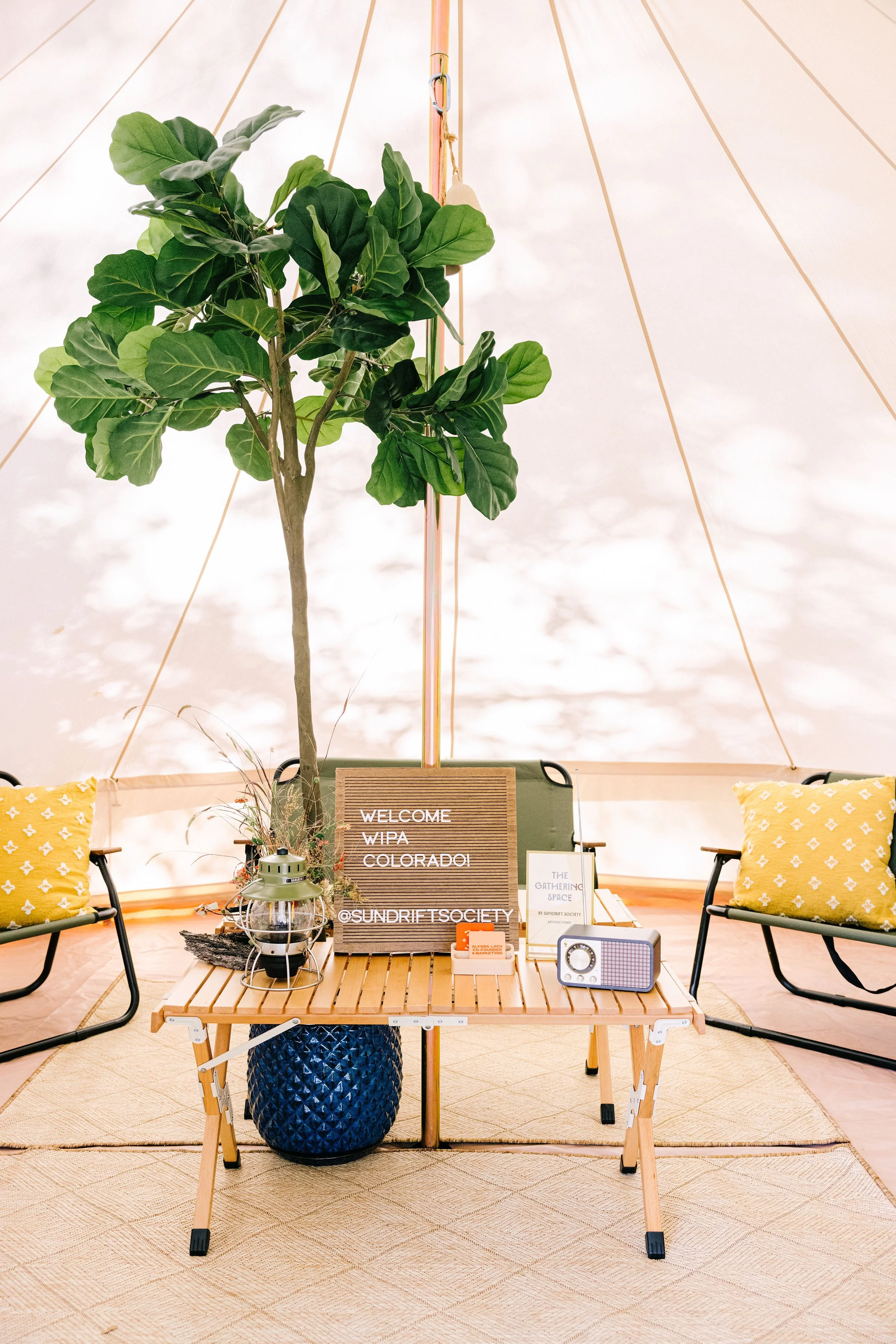 Indoor gathering space with a large green potted plant, a wooden table, yellow cushions, a welcome sign, and vintage-style radio, all under a tent-like ceiling with soft lighting.
