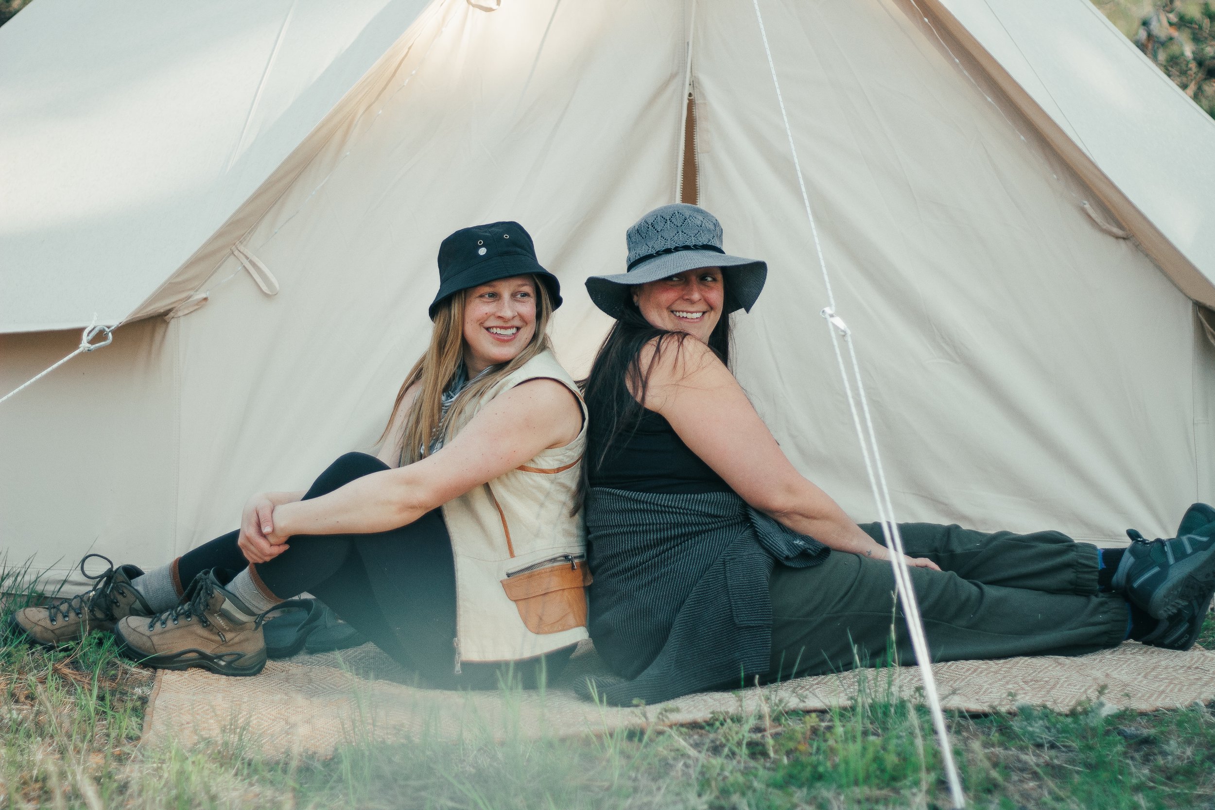 Two women sitting back-to-back under a large camping tent, smiling and enjoying outdoor camping. They wear casual outdoor clothing, including hats, and are seated on a mat on grassy ground.