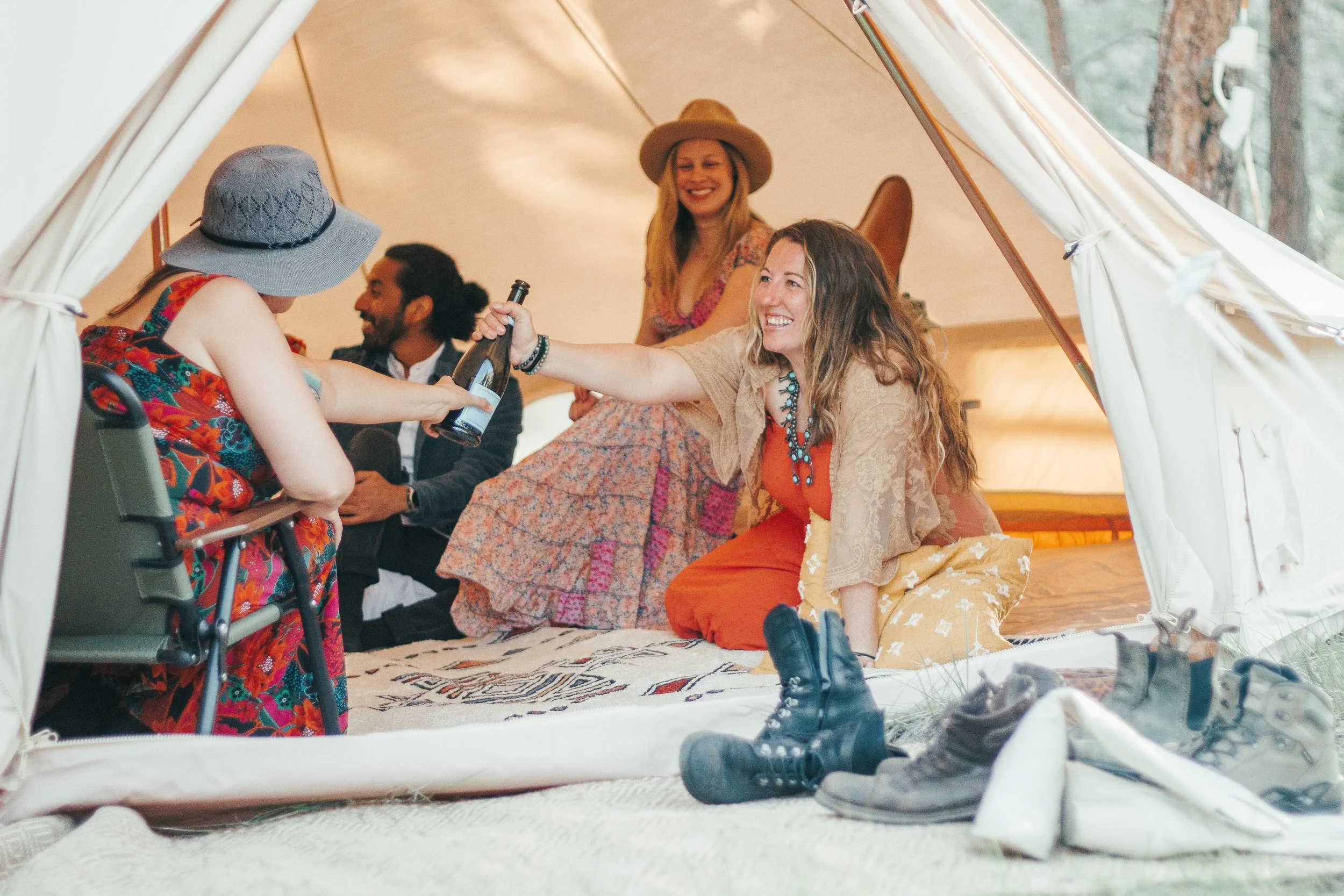 Group of friends inside a tent at a campsite, smiling and having fun. One woman in beige and orange clothes sits on the ground, another woman in floral dress and hat is smiling, a woman in a patterned dress and sun hat is laughing, and a man with a b