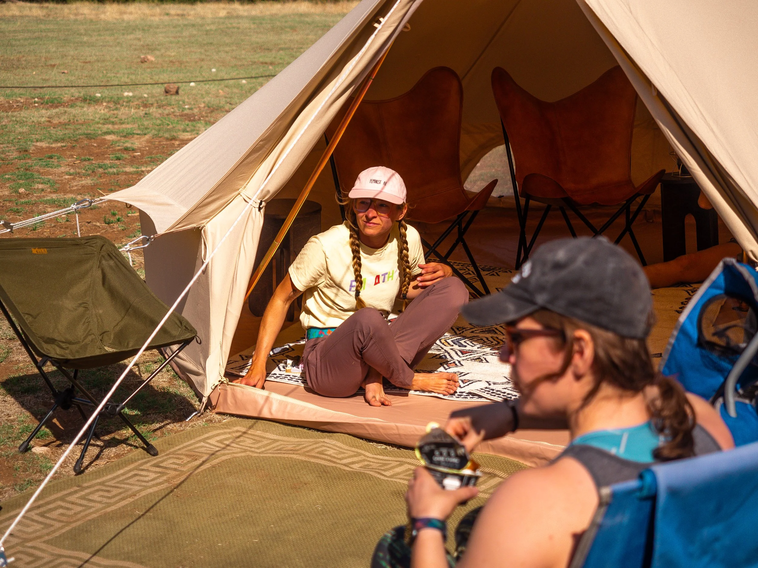 A woman with long braided hair, wearing glasses, a pink cap with text, and a white T-shirt sitting on a patterned rug inside a canvas tent during daytime. Several chairs are visible inside the tent, and other people are seated nearby outside the tent