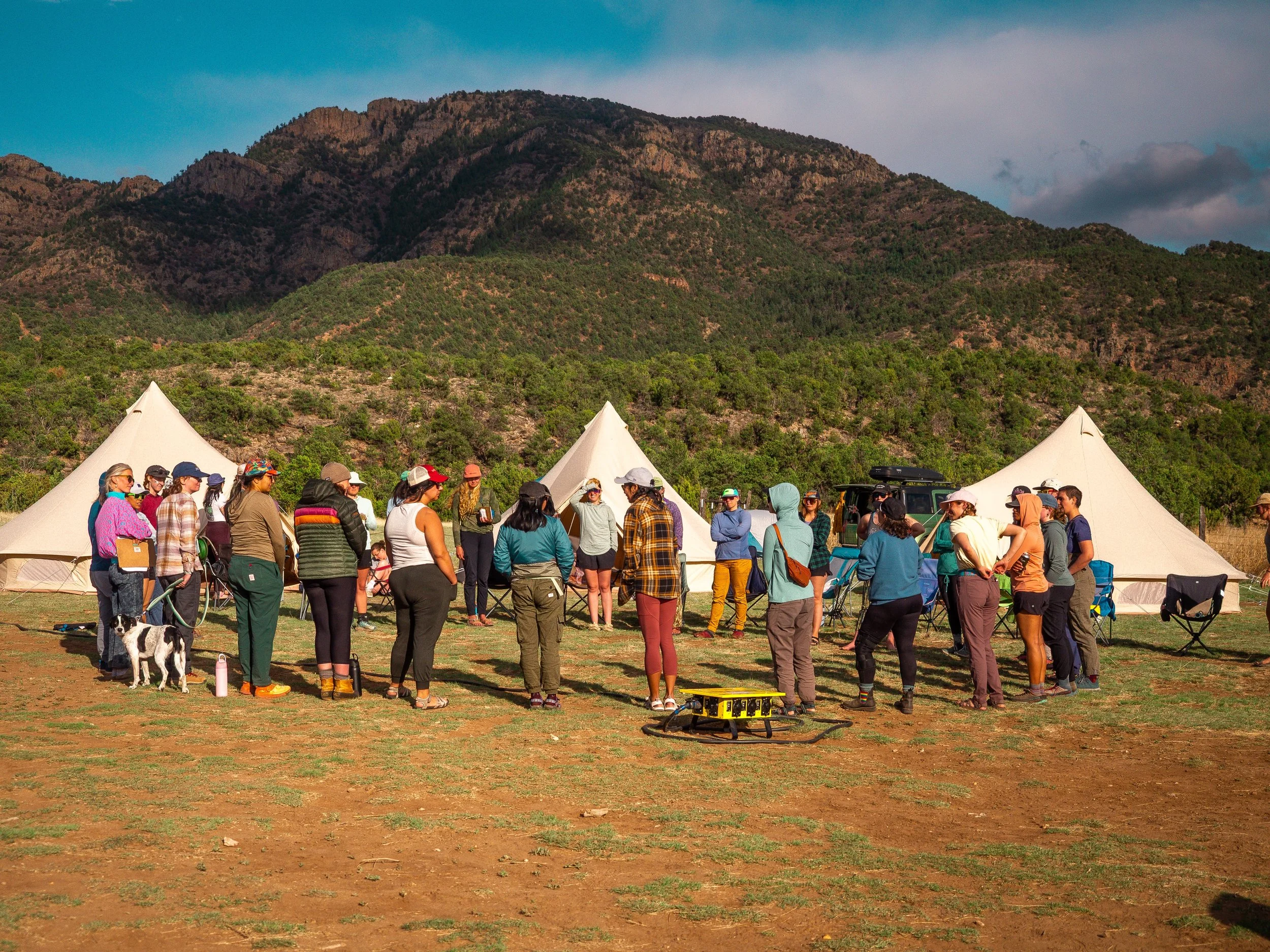 Group of people gathered outdoors near white tents with a mountain range in the background, during daytime.