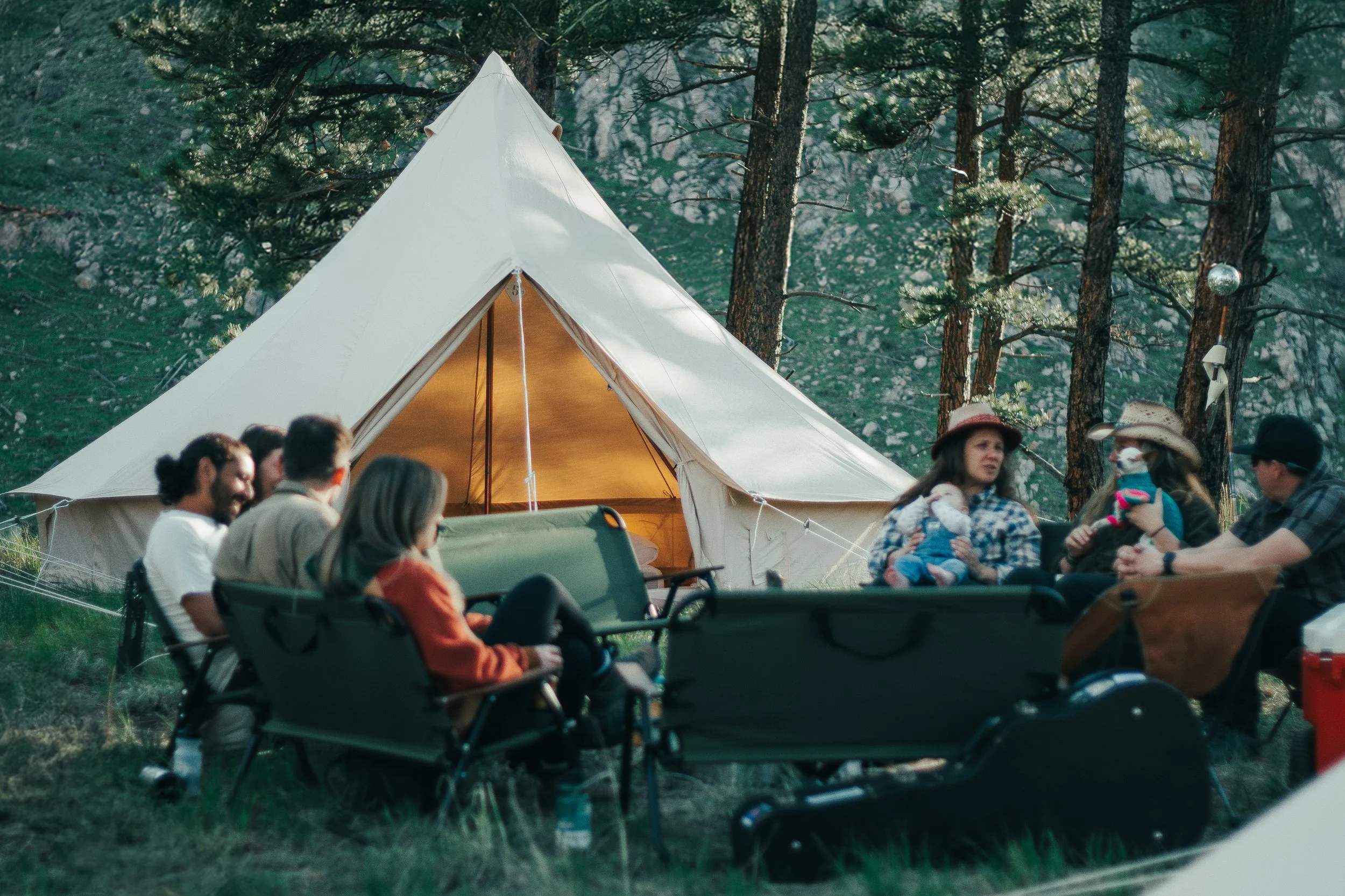 Group of people sitting around a campfire near a large white tipi in a forested area with trees and rocky terrain visible in the background.