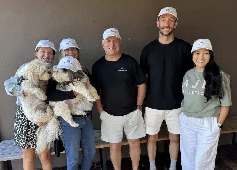 Group of five people, three women and two men, standing together and smiling, wearing white caps. Two women are holding light-colored dogs, and the women and one man are dressed casually in shorts and t-shirts.
