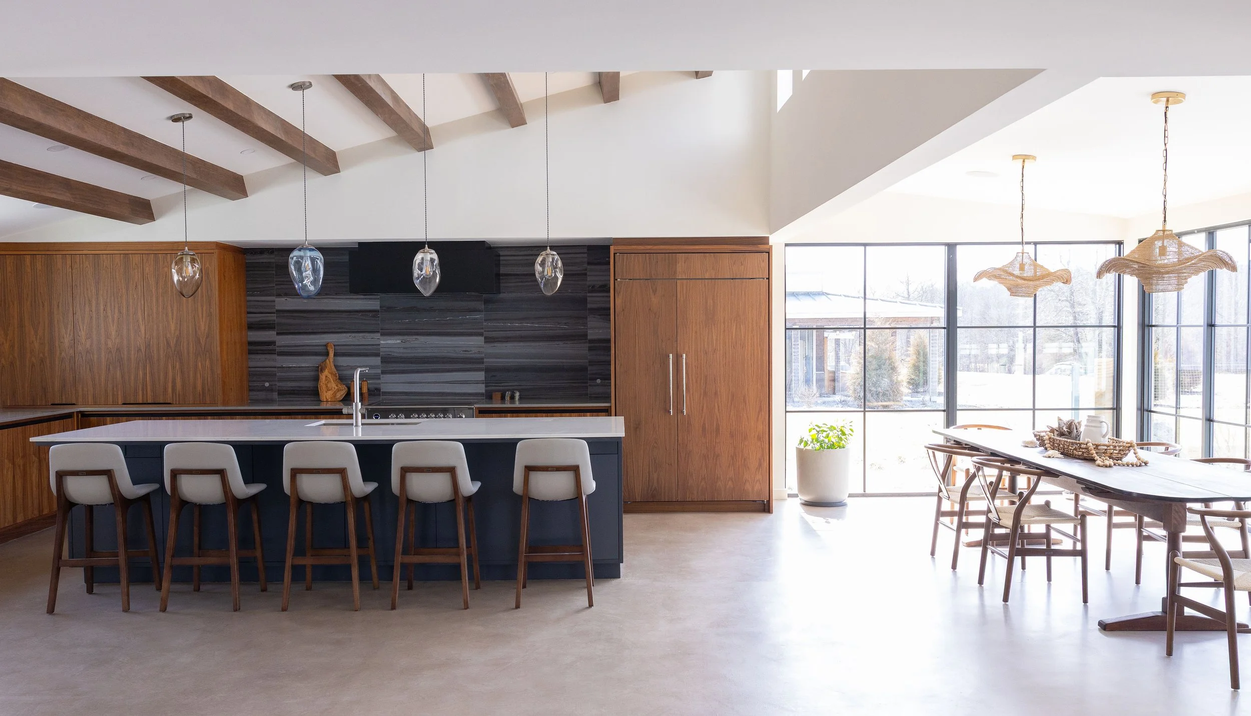 Modern dining and kitchen area with large windows, wooden beams, a kitchen island with chairs, and hanging pendant lights.