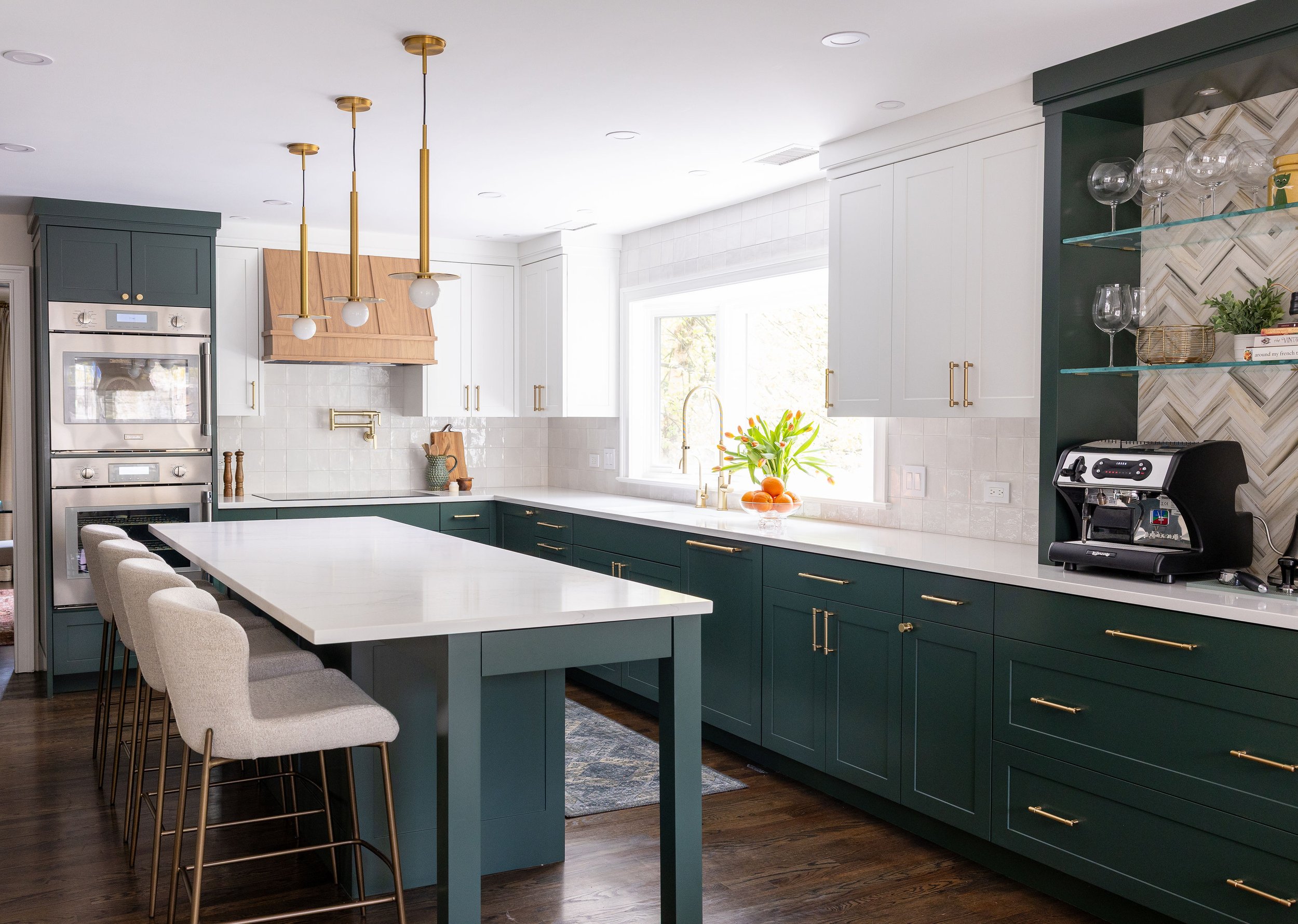Modern kitchen with dark green cabinets, white countertops, gold hardware, and a white tile backsplash. Contains a kitchen island, wall of windows, pendant lights, built-in oven, coffee machine, and open shelving with glassware and plants.