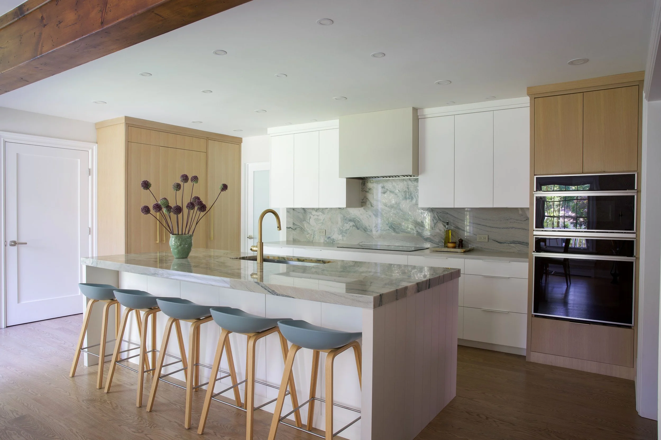Modern kitchen with white cabinets, marble backsplash, island with marble countertop, blue barstools, and a vase with purple flowers