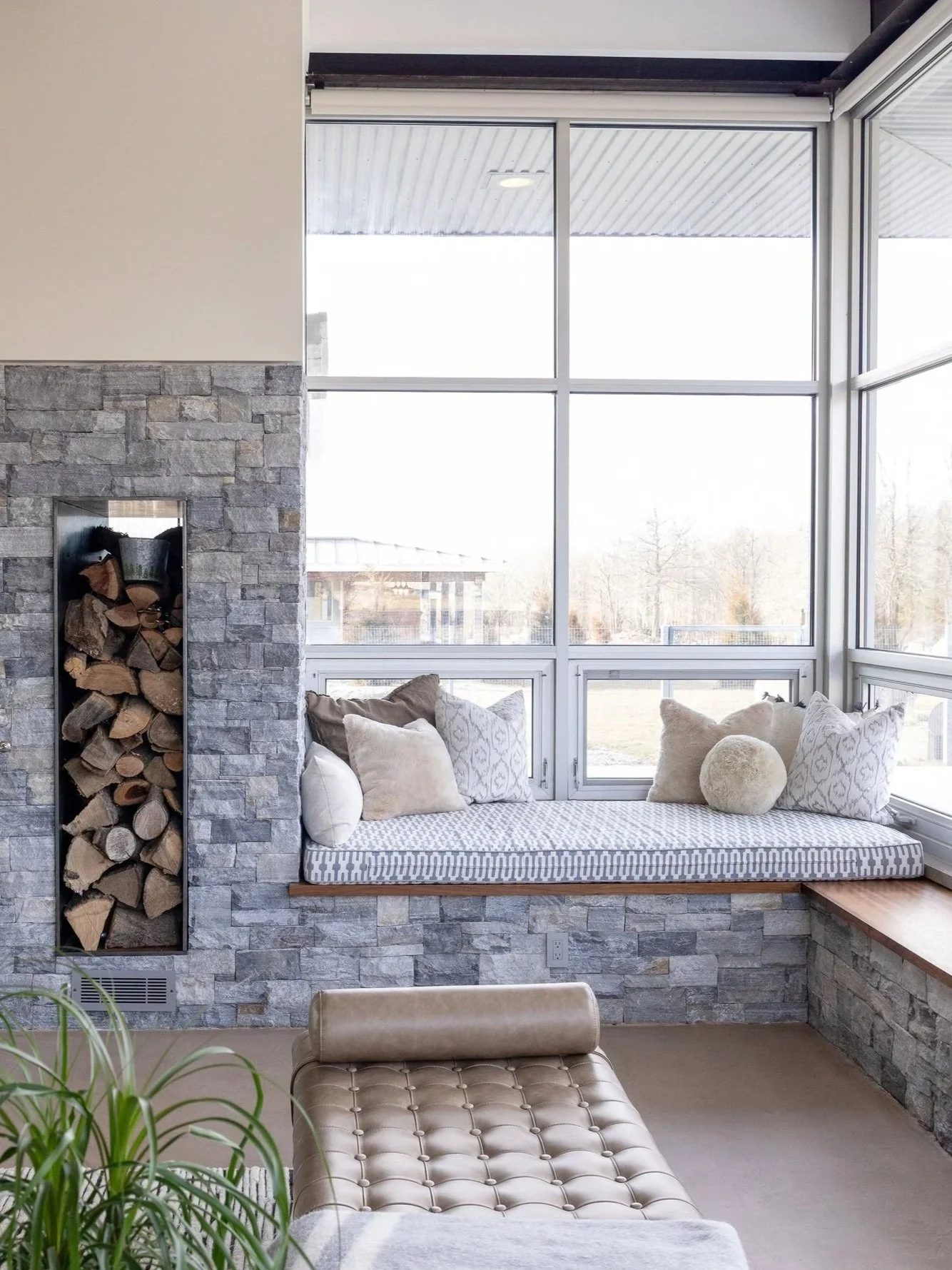 Sunroom with large windows, a built-in window seat with beige and patterned cushions, a wood-burning firewood storage, a beige tufted chaise lounge, and a potted houseplant.