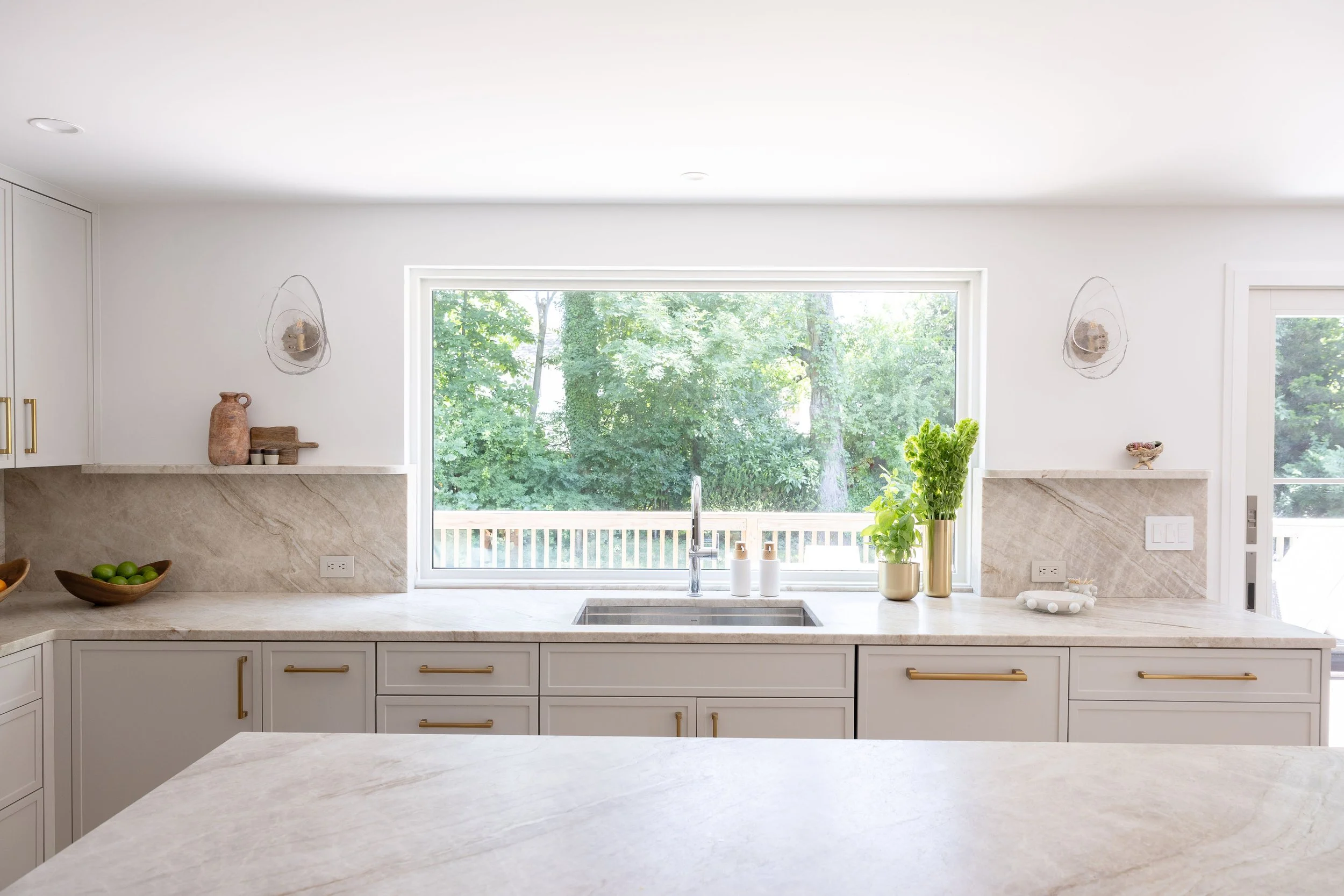 Bright modern kitchen with large window overlooking trees, white cabinets with gold handles, marble countertops, and minimal decor.