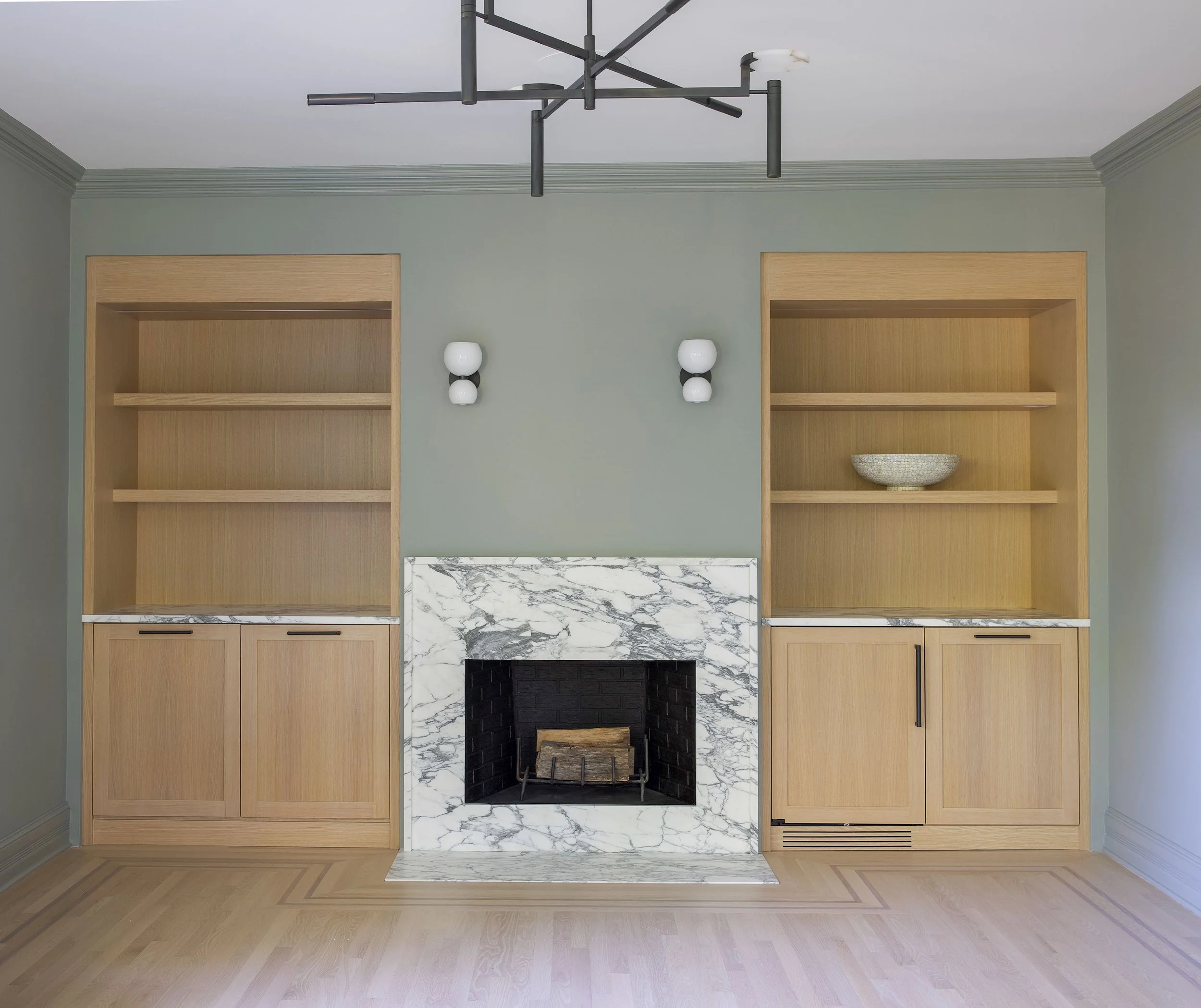 Living room with built-in wooden bookshelves on each side of a marble fireplace, light green walls, and modern black and white wall sconces.