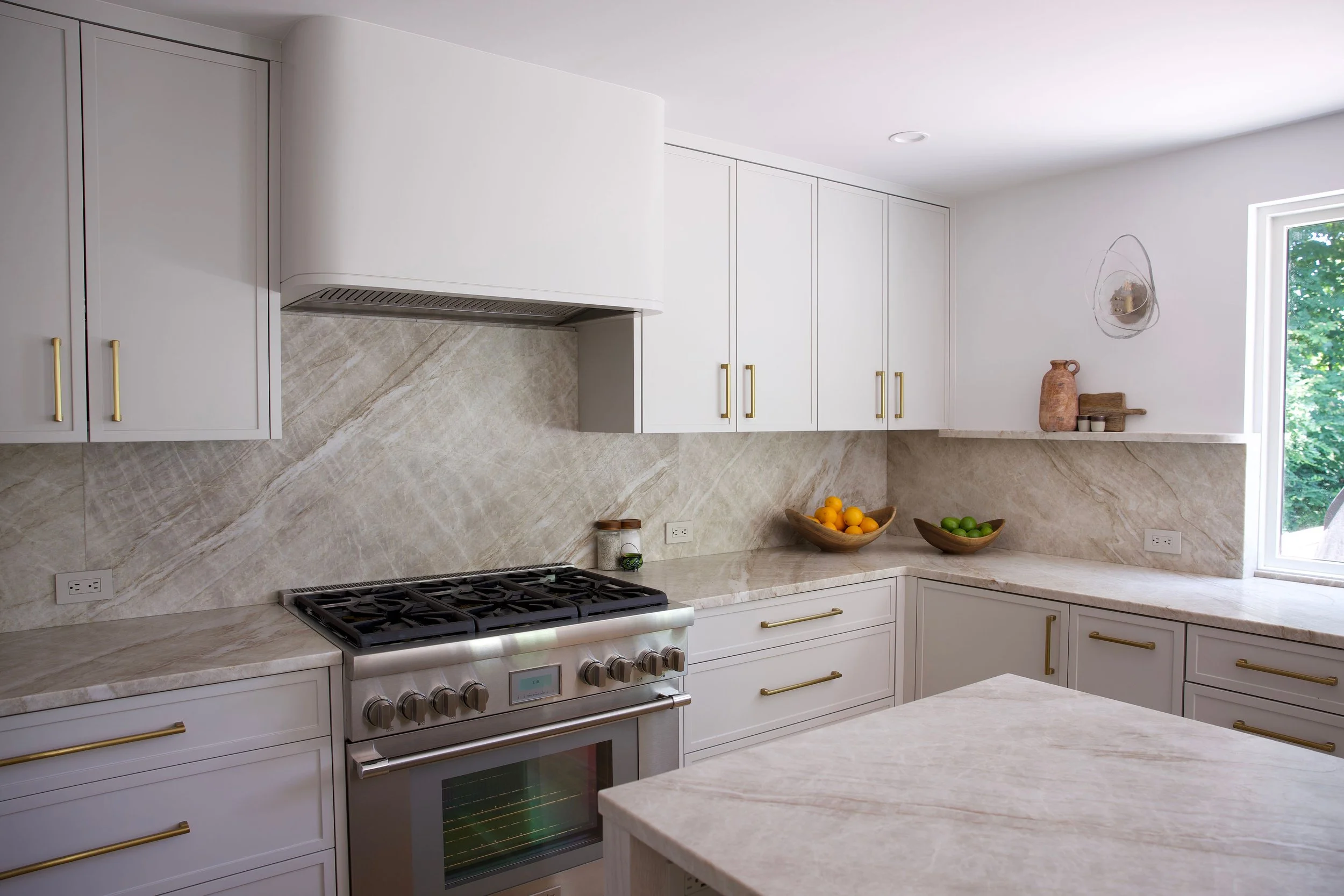 Modern kitchen with white cabinetry, gold handles, marble countertops, a stovetop, and a window with green trees outside.