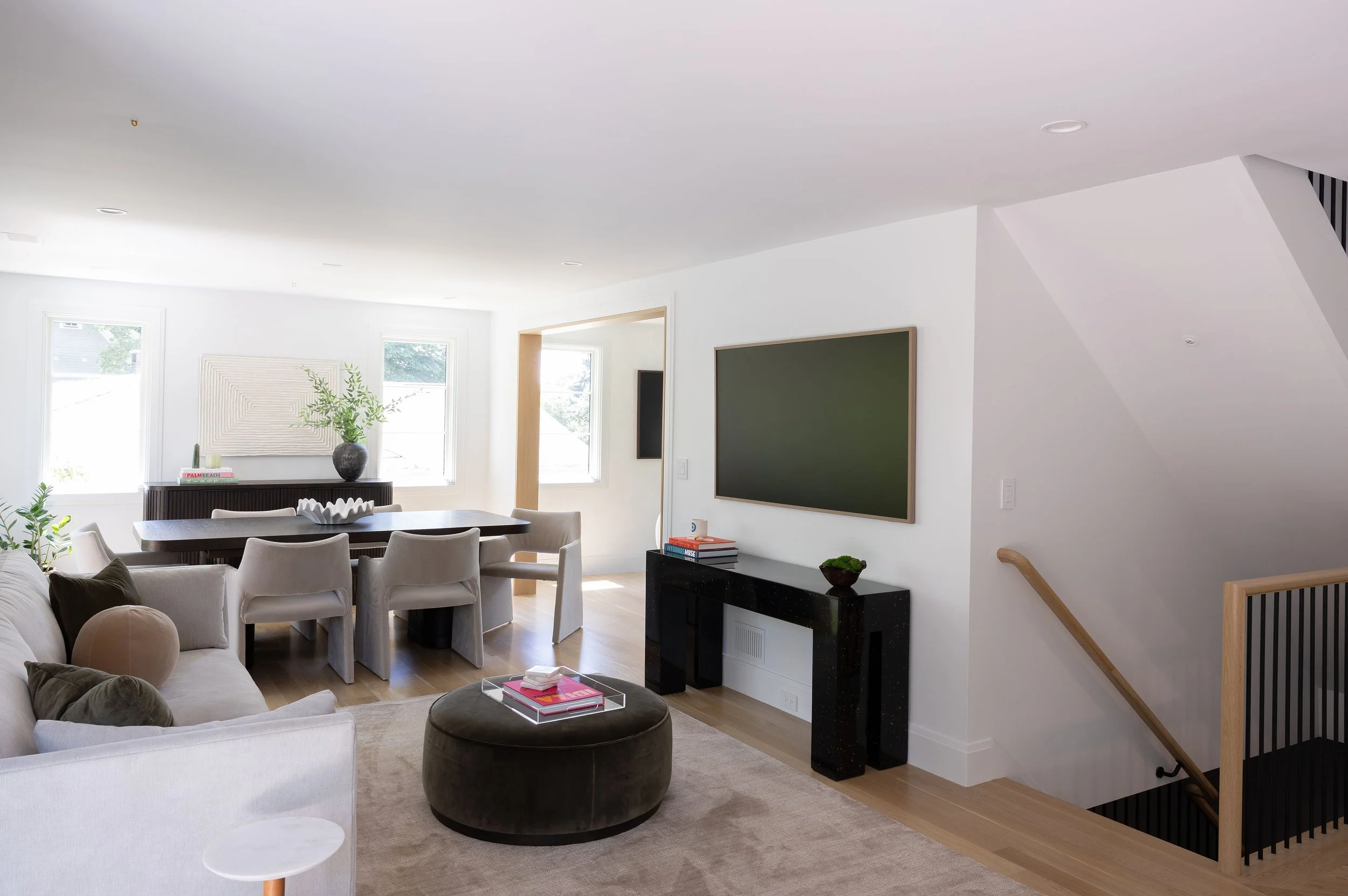 Modern living room with white walls, a black TV on a black console, a beige sectional sofa with pillows, a dark ottoman with trays, and a wooden staircase with black slats. Large windows let in natural light.