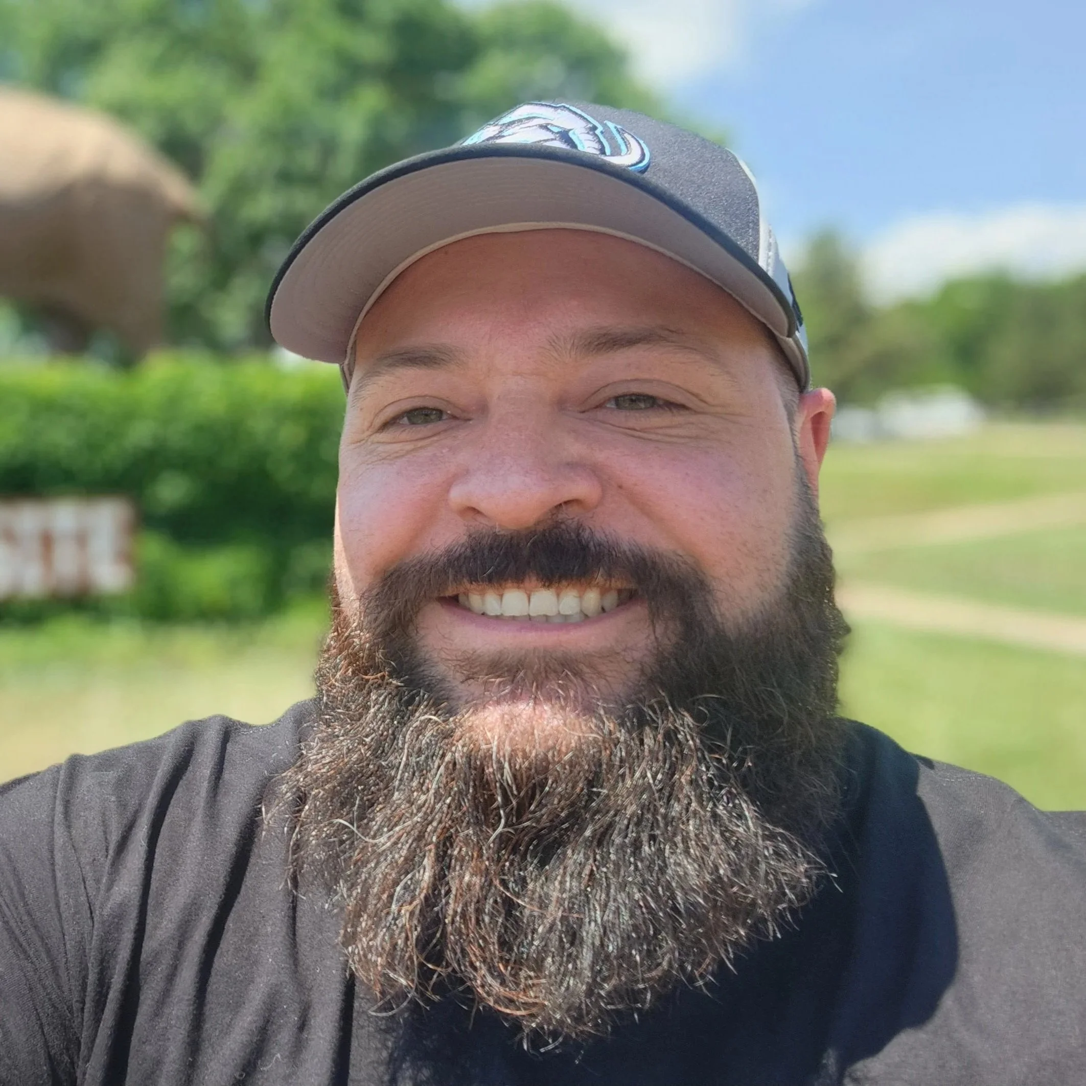 Close-up of a smiling man wearing a baseball cap outdoors, with trees and blue sky in the background.