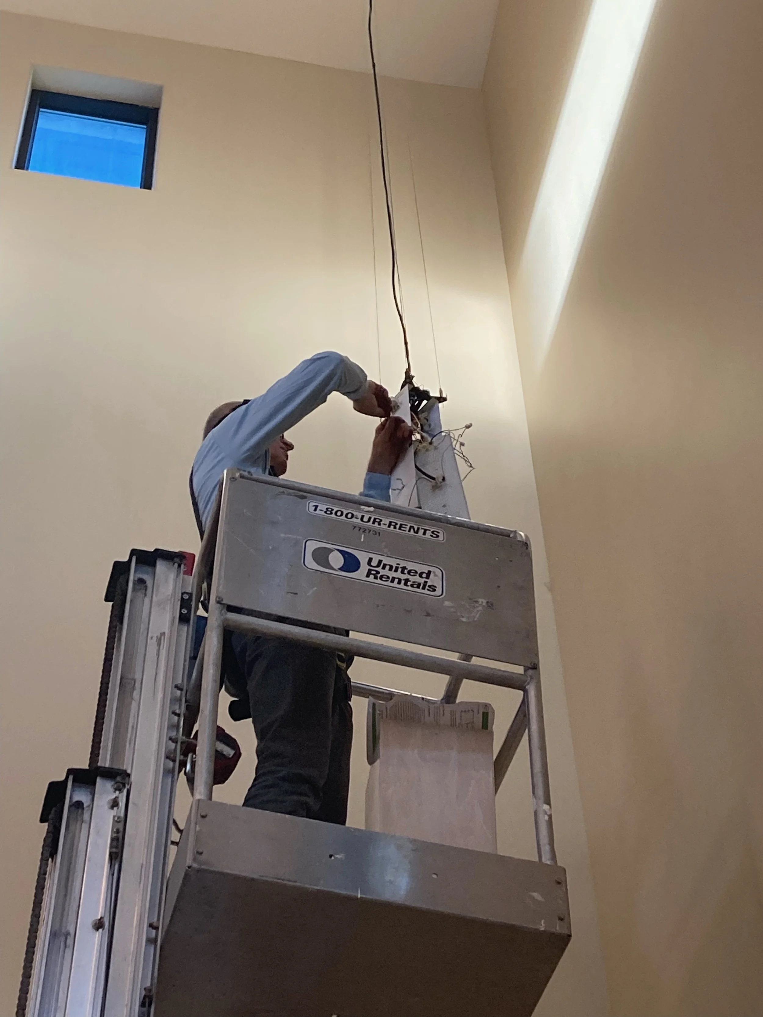 A person working on electrical wiring while standing on a lift inside a room with cream-colored walls and a high ceiling.