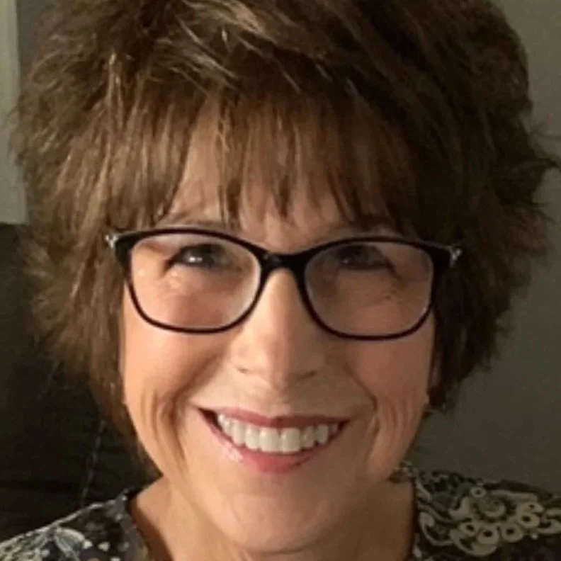Close-up of a smiling woman with short curly hair, wearing glasses and a patterned top.