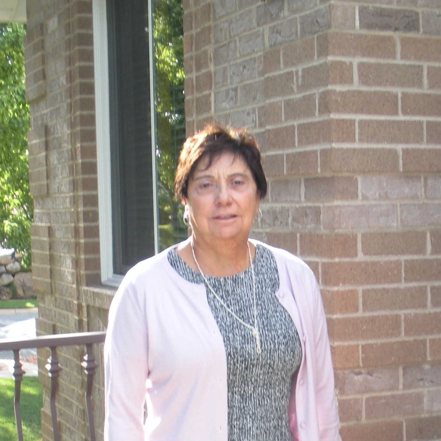 A woman with short dark hair and earrings standing outside near a brick building, wearing a patterned top and a light pink cardigan, with a necklace.
