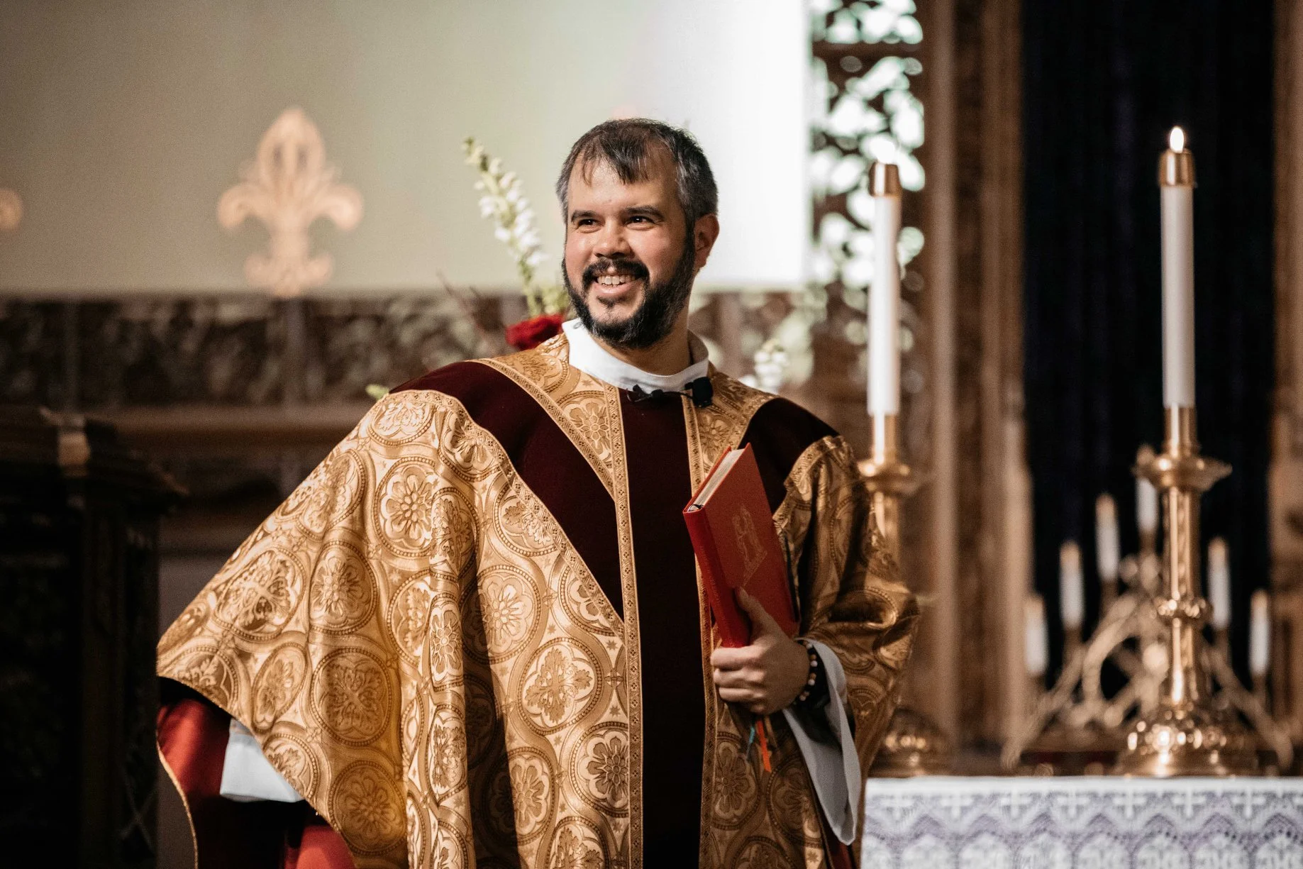 A man dressed in ornate ceremonial robes holding a red book, smiling inside a church with lit candles on gold holders in the background.