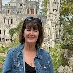 A woman with dark hair and sunglasses on her head, wearing a denim jacket, standing outdoors in front of a castle-like structure and greenery.