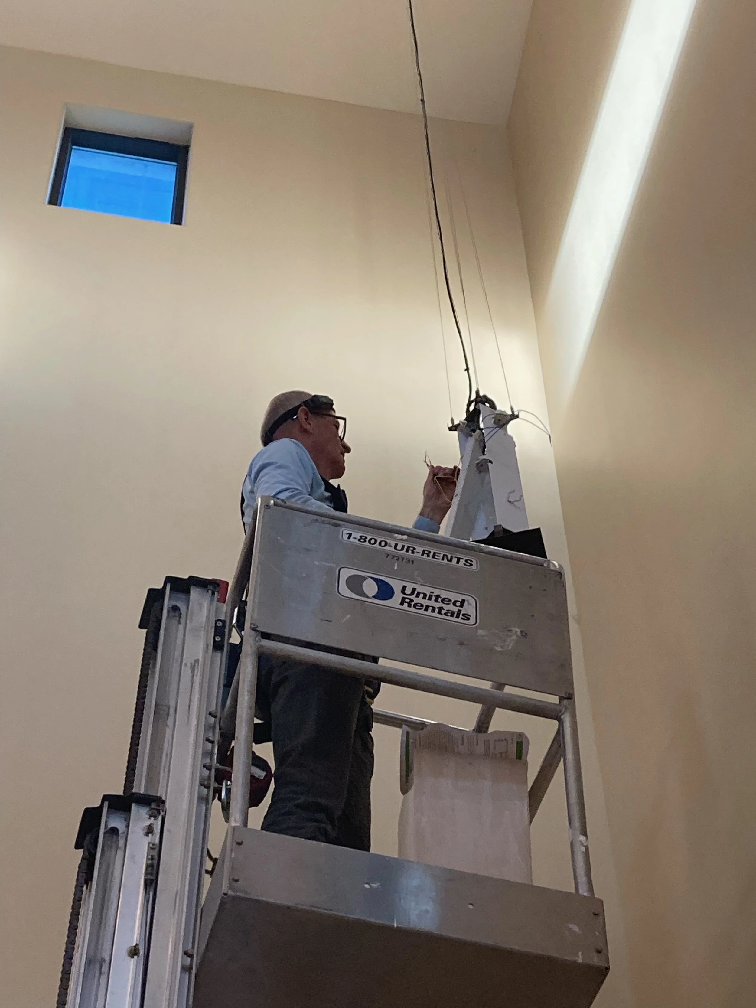 A person in safety glasses and a hat working on electrical wiring on a high ceiling, standing on a mobile lift labeled 'United Rentals' inside a room with beige walls and a small square window.