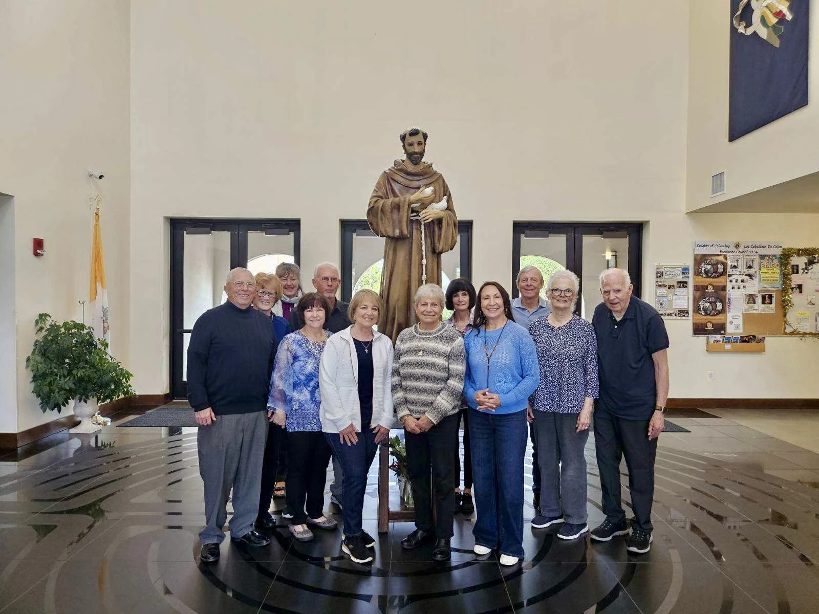 Group of eleven people, mostly women and two men, standing in front of a large religious statue of a saint holding a cross, inside a church or community center with plaques and banners on the walls.