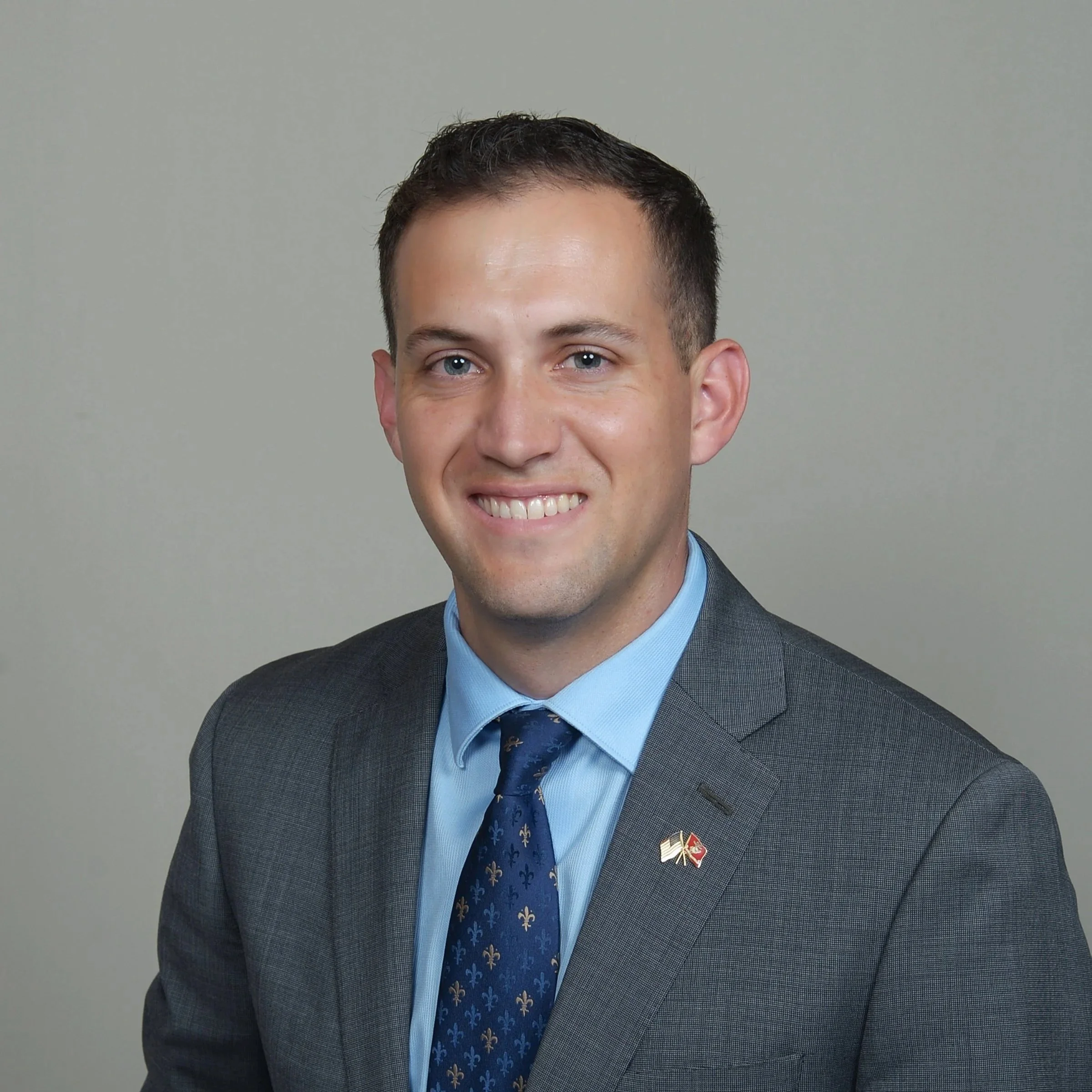 Photograph of a smiling man in a dark gray suit with a blue shirt, wearing a patterned blue tie and a pin on his lapel, posing against a plain gray background.