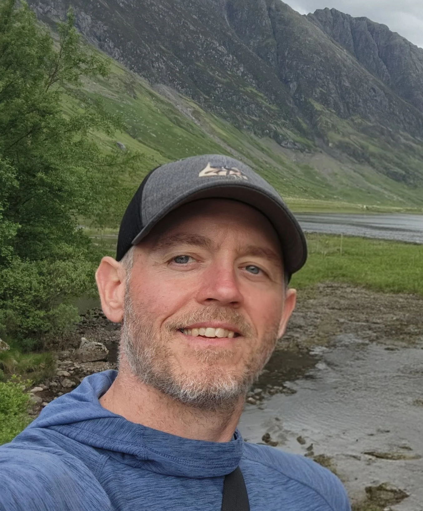 A man taking a selfie outdoors near a river, with mountains and green hills in the background.