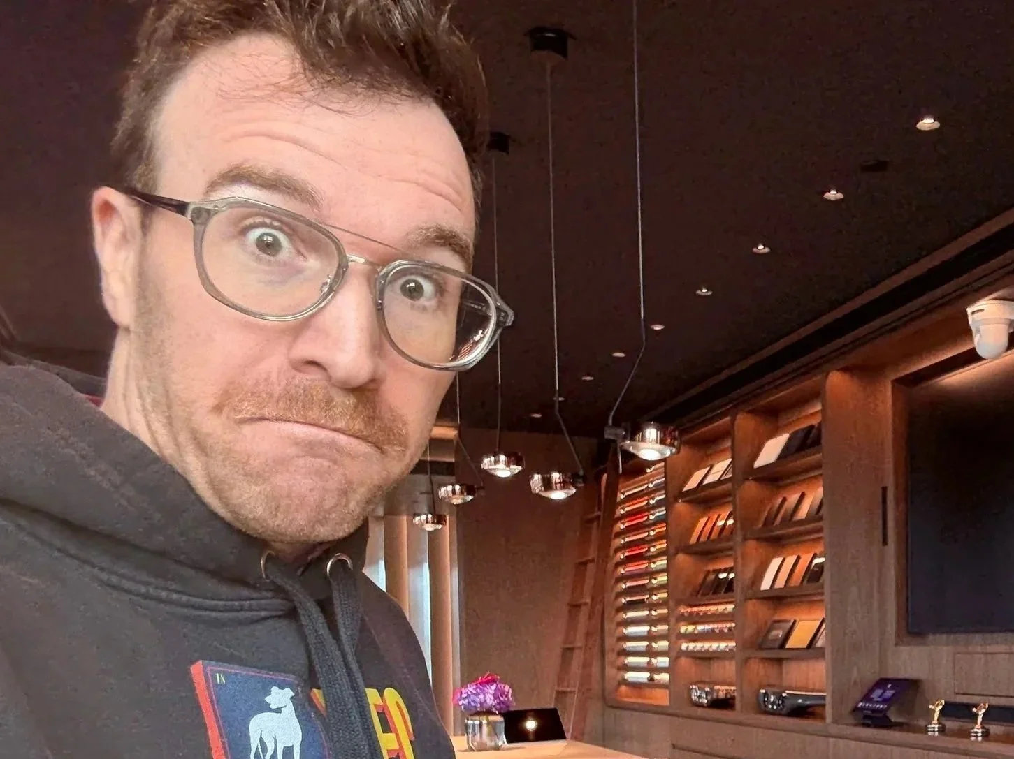 A man wearing glasses and a dark hoodie with colorful text stands in a modern, wood-paneled retail store with sunglasses displayed on shelves behind him.