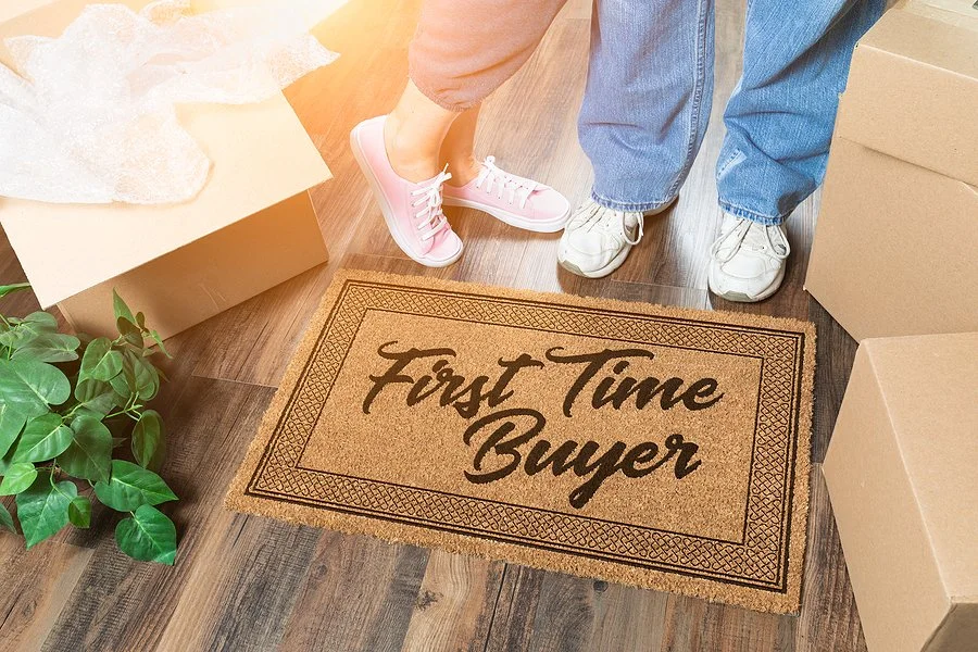 Close-up of a doormat with the words "First Time Buyer" in front of a couple with boxes, suggesting moving into a new home.