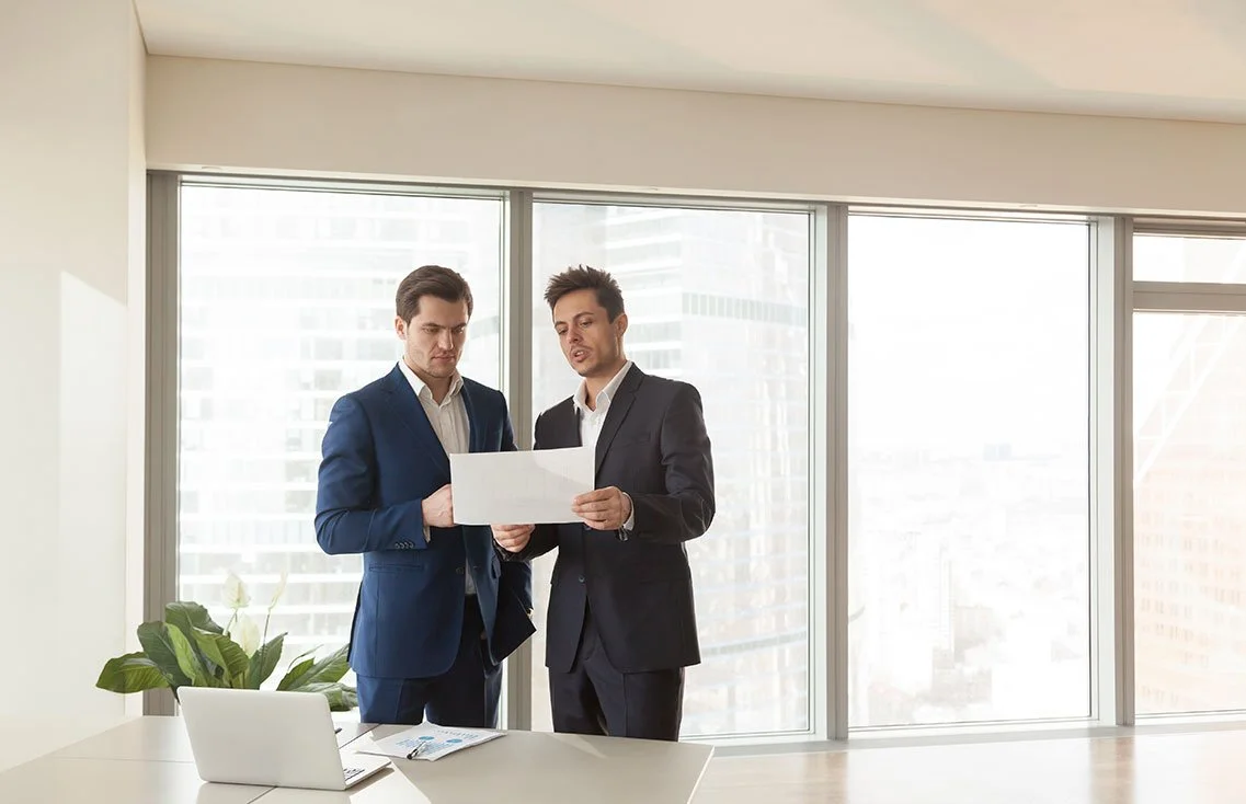 Two businessmen in suits having a discussion while looking at a document in a modern office with large windows and a city view.