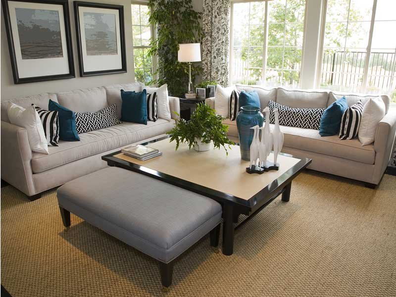 Living room with two white sofas, a light gray ottoman, a black-framed coffee table with decorative vases and a plant, and large windows with curtains and greenery outside.